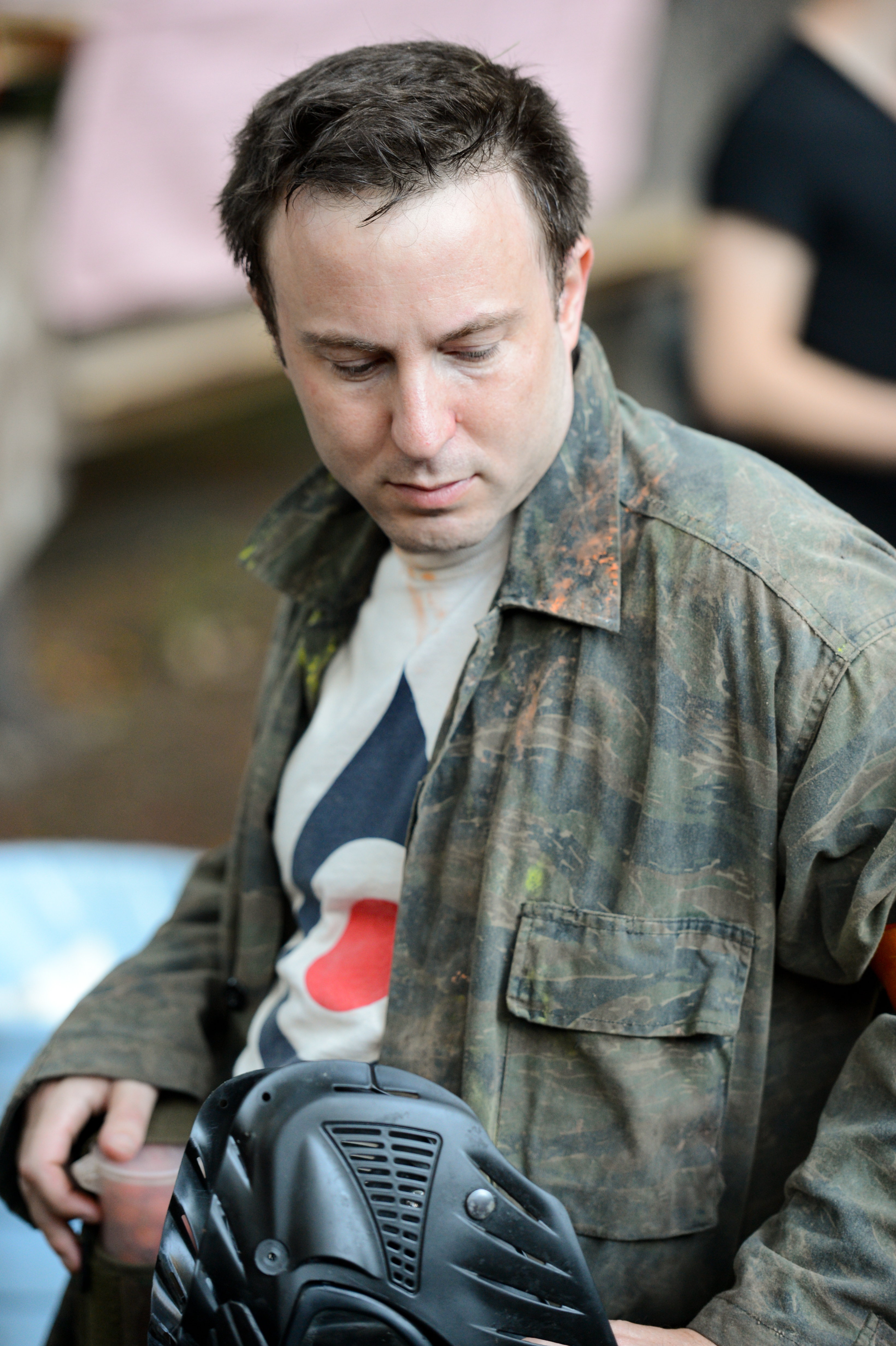 A person wearing a camouflage jacket holds a black paintball mask while looking down.