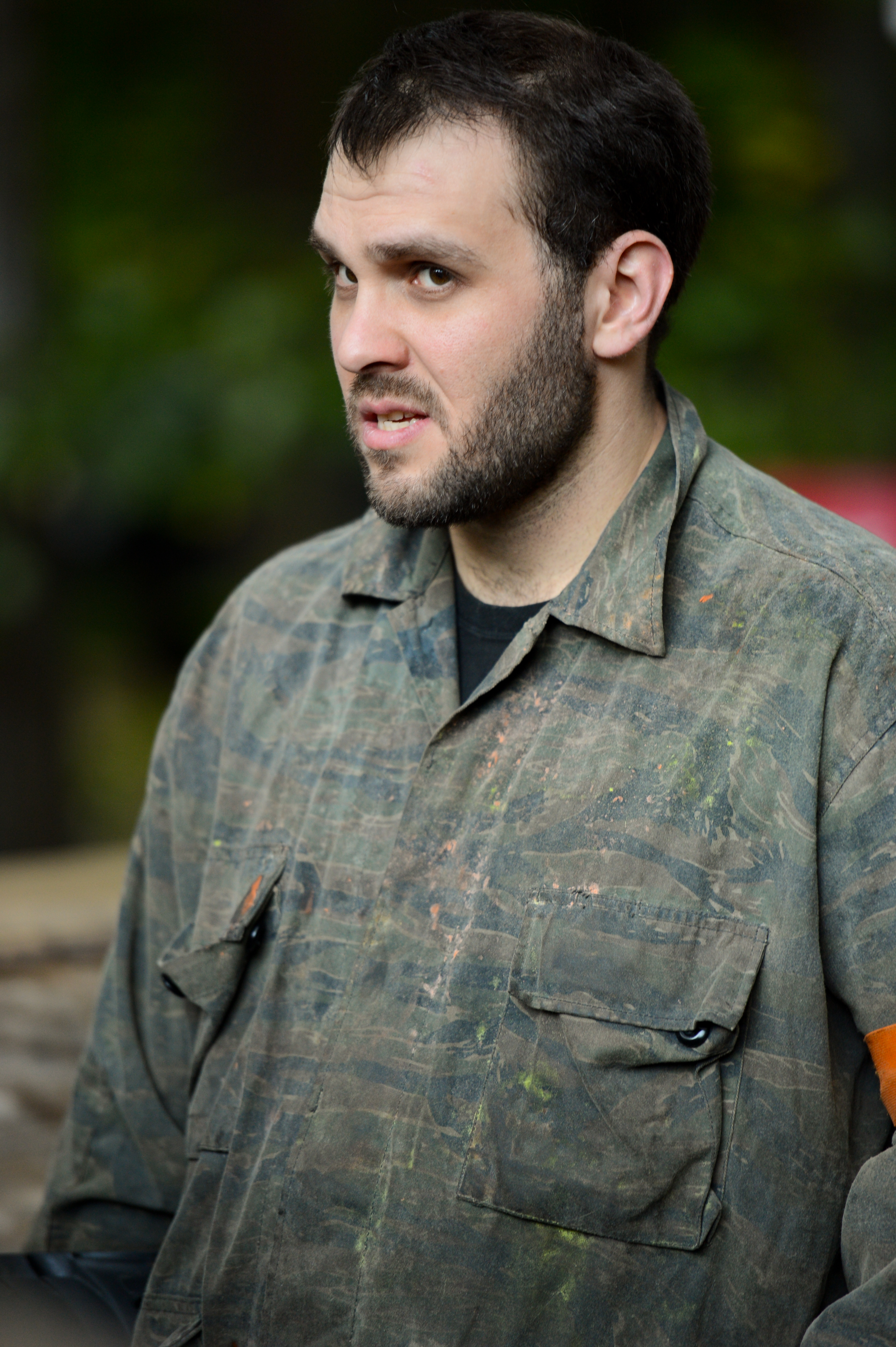 A man in a camouflage jumpsuit with paint stains looks to the side with a focused expression.