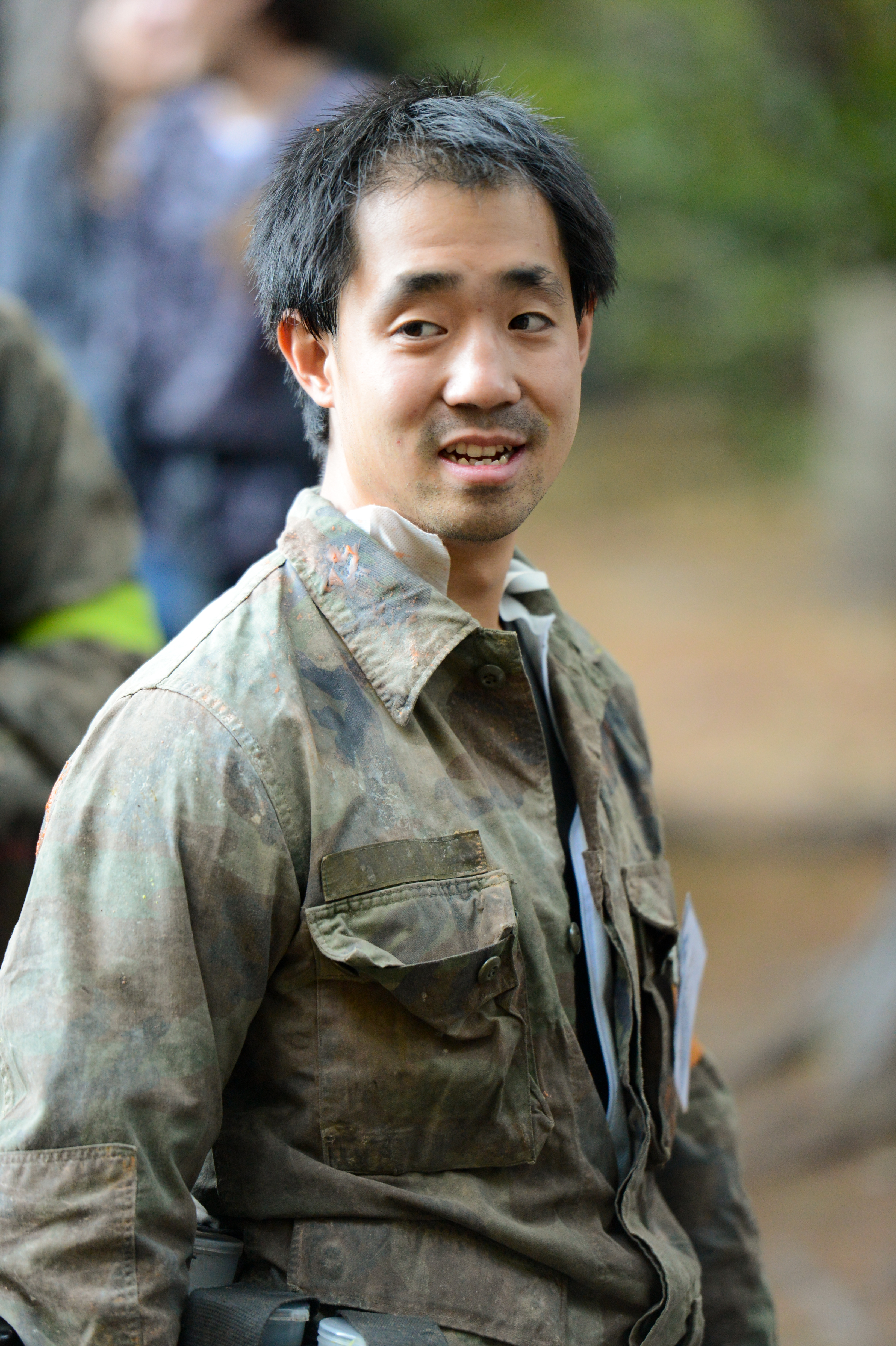 A person wearing a camouflage jacket with paint splatters looks to the side in an outdoor setting.