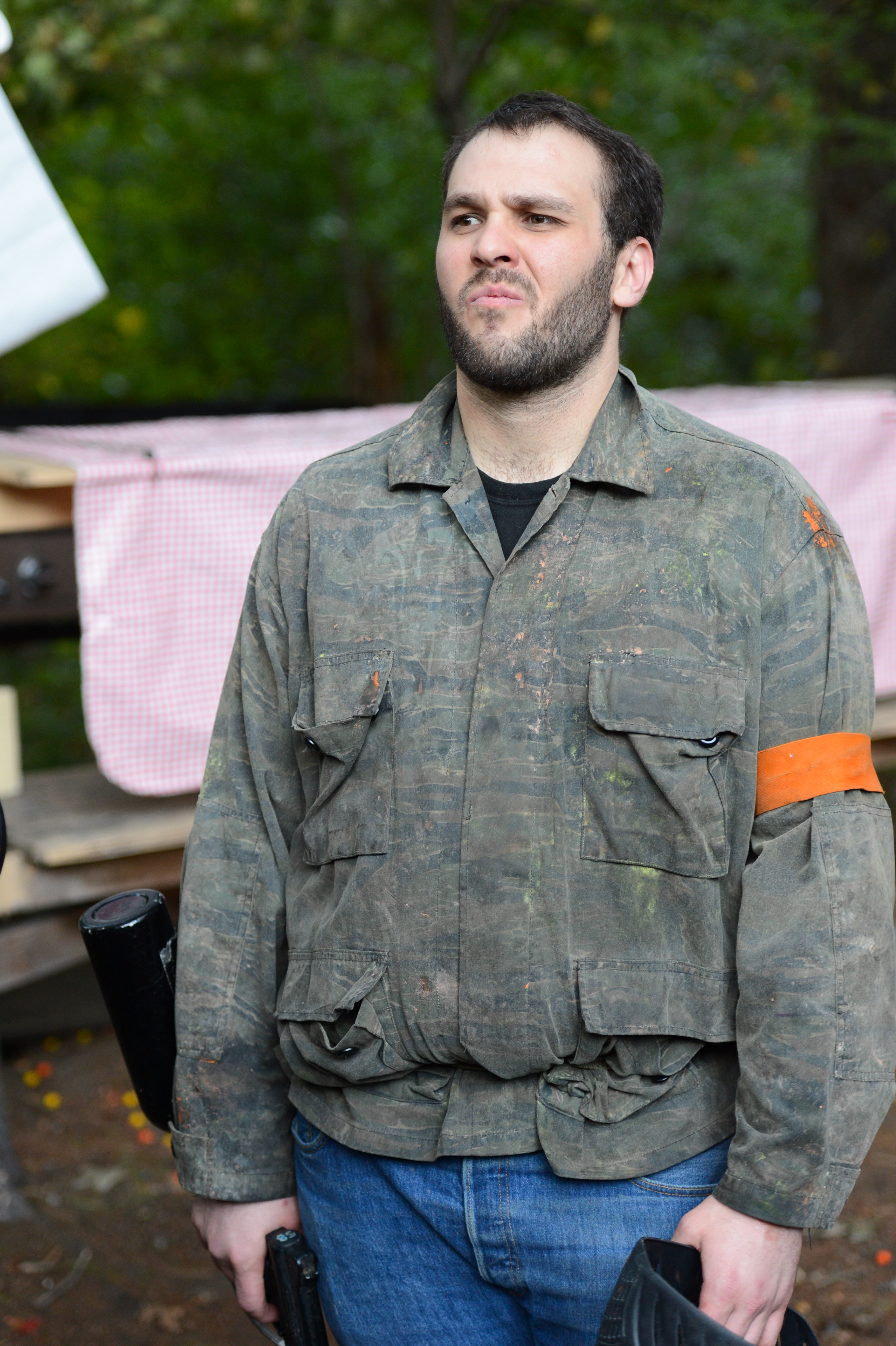A person in a camouflage jacket with an orange armband holds paintball gear and looks off to the side.