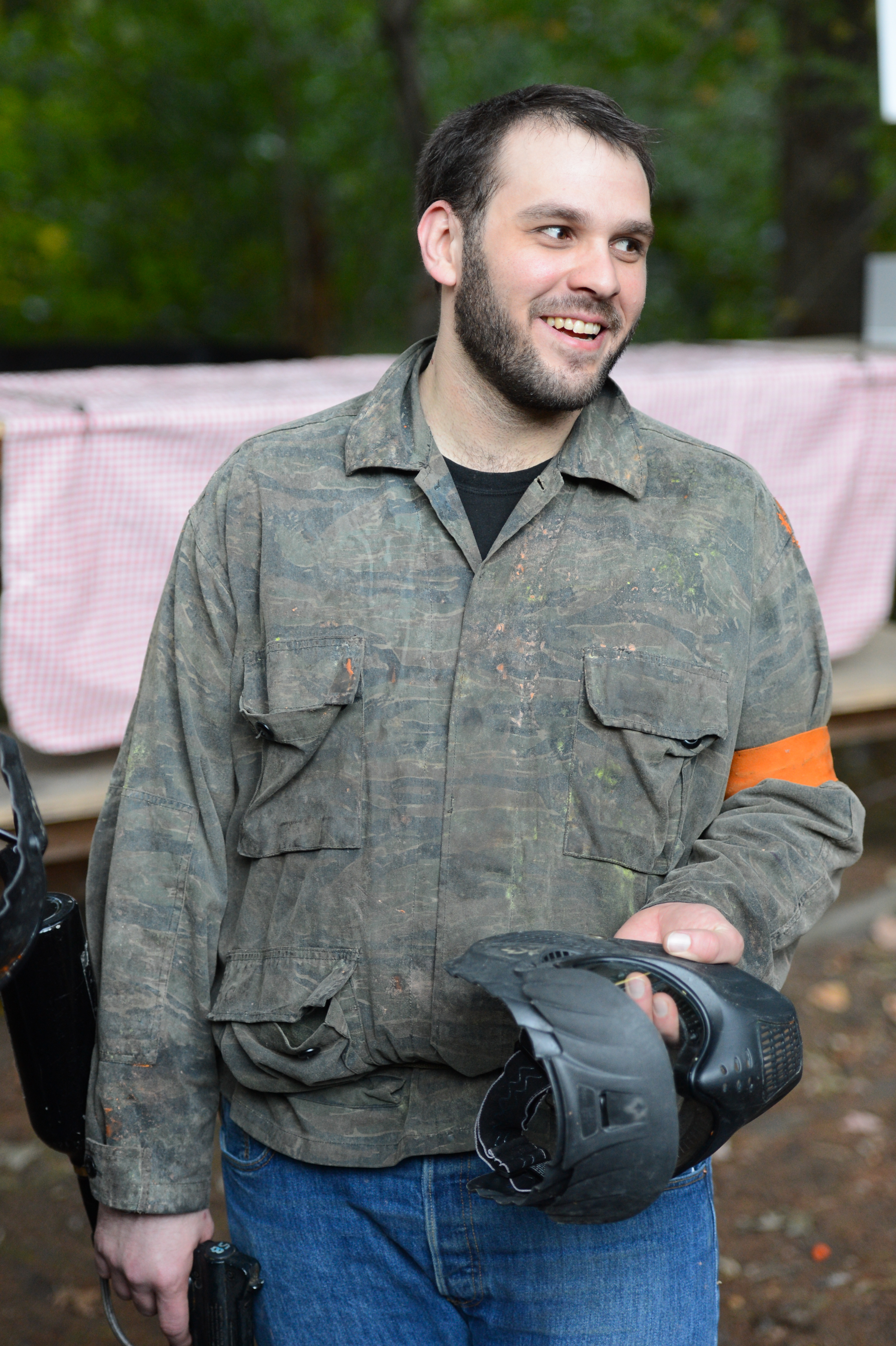 A man in a camouflage jacket smiles while holding a paintball mask and gear outdoors.