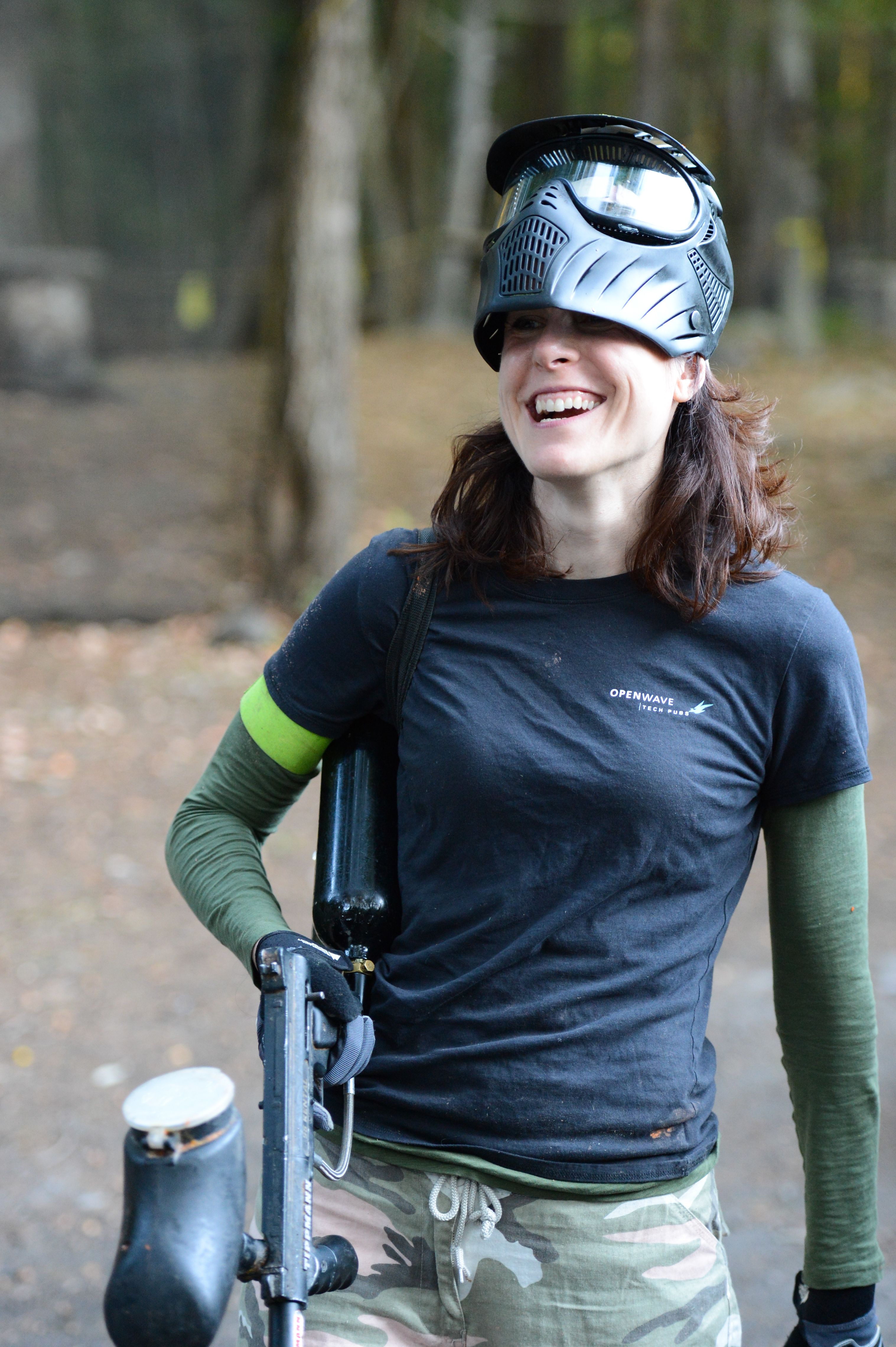 A person wearing a paintball mask and camouflage pants smiles while holding a paintball gun in an outdoor setting.