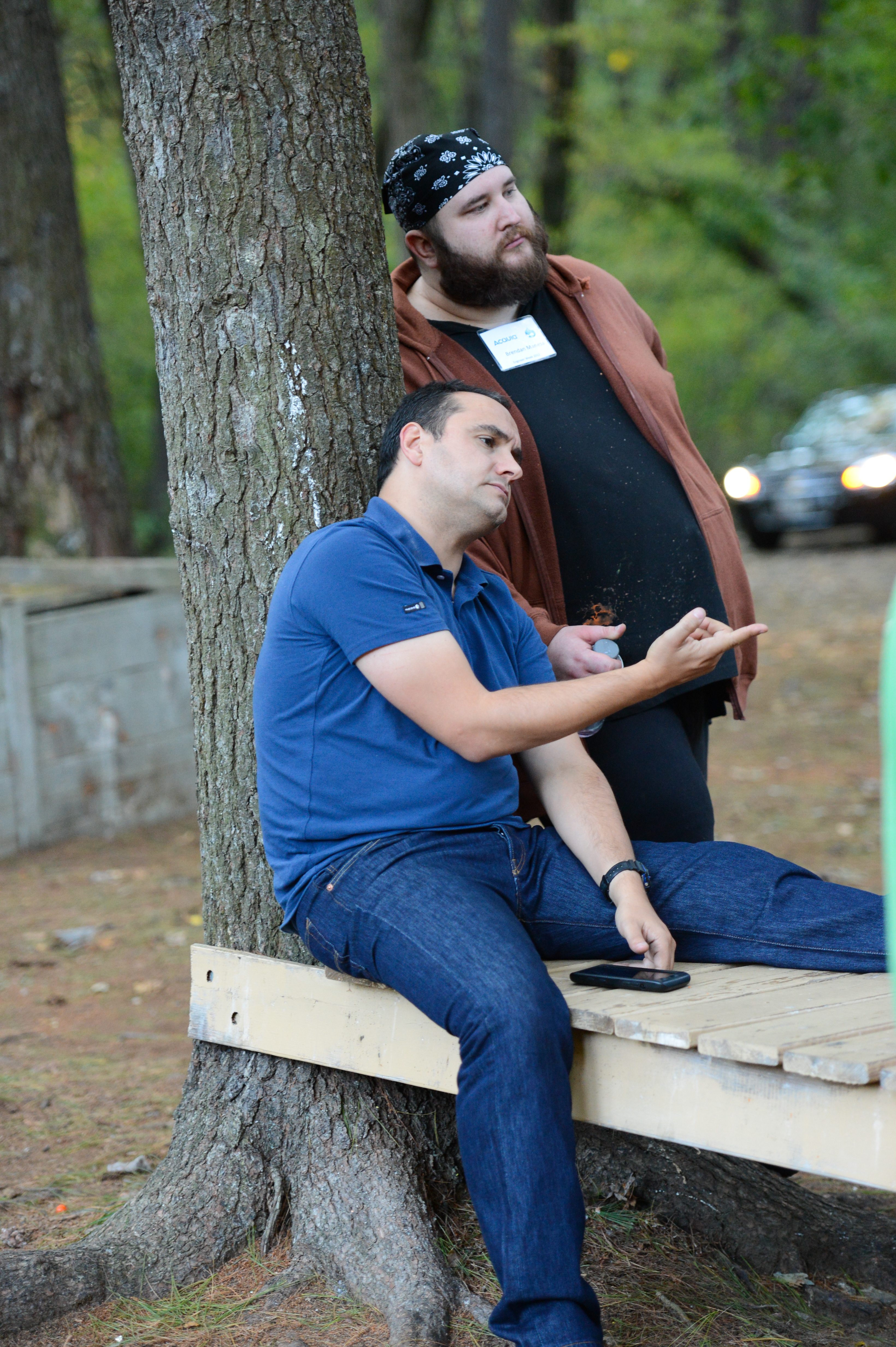 Two men outdoors, one sitting on a wooden bench gesturing while the other leans against a tree listening.