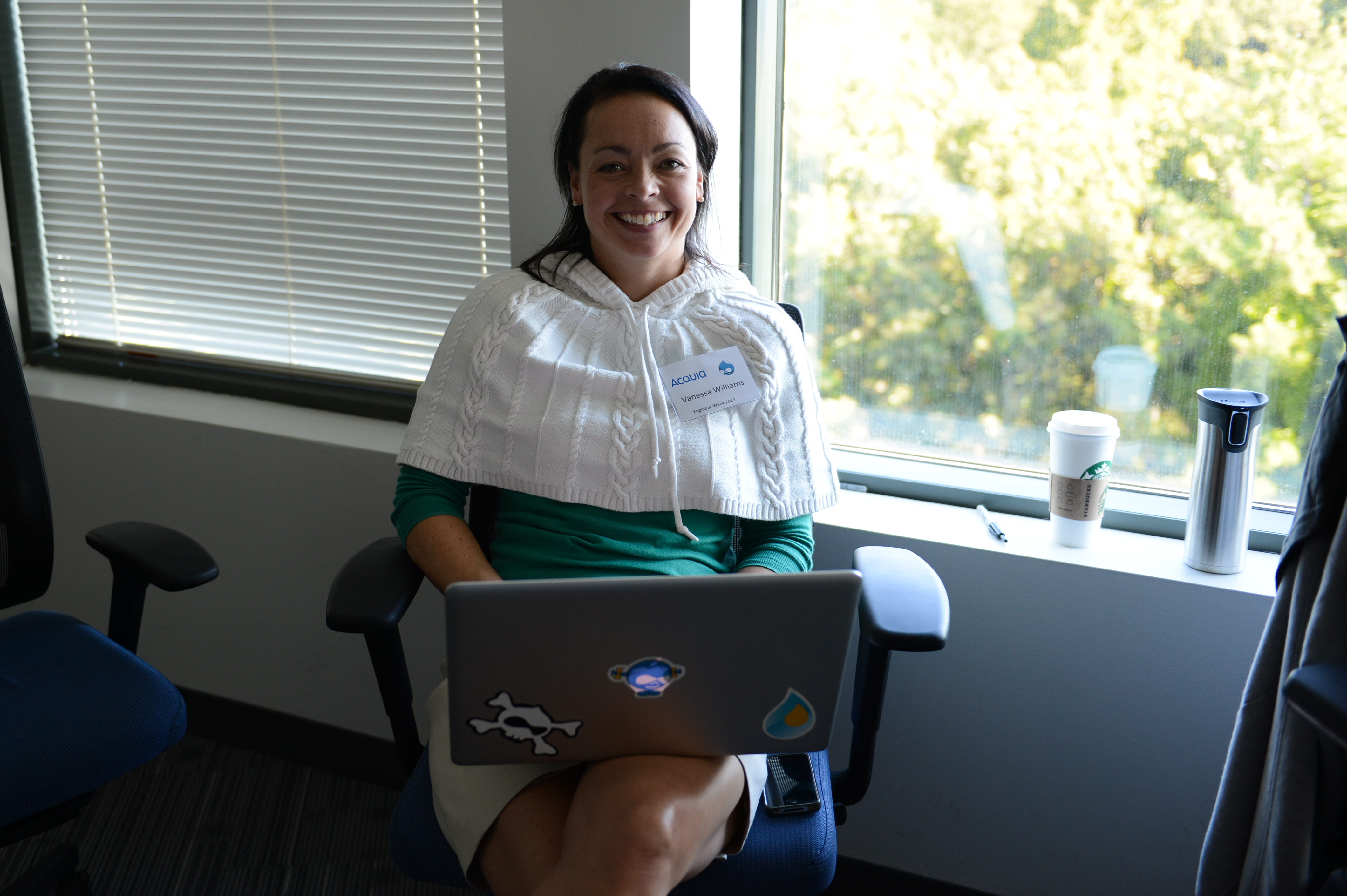 A person sits in an office chair, smiling while using a laptop, with a name tag on their sweater.