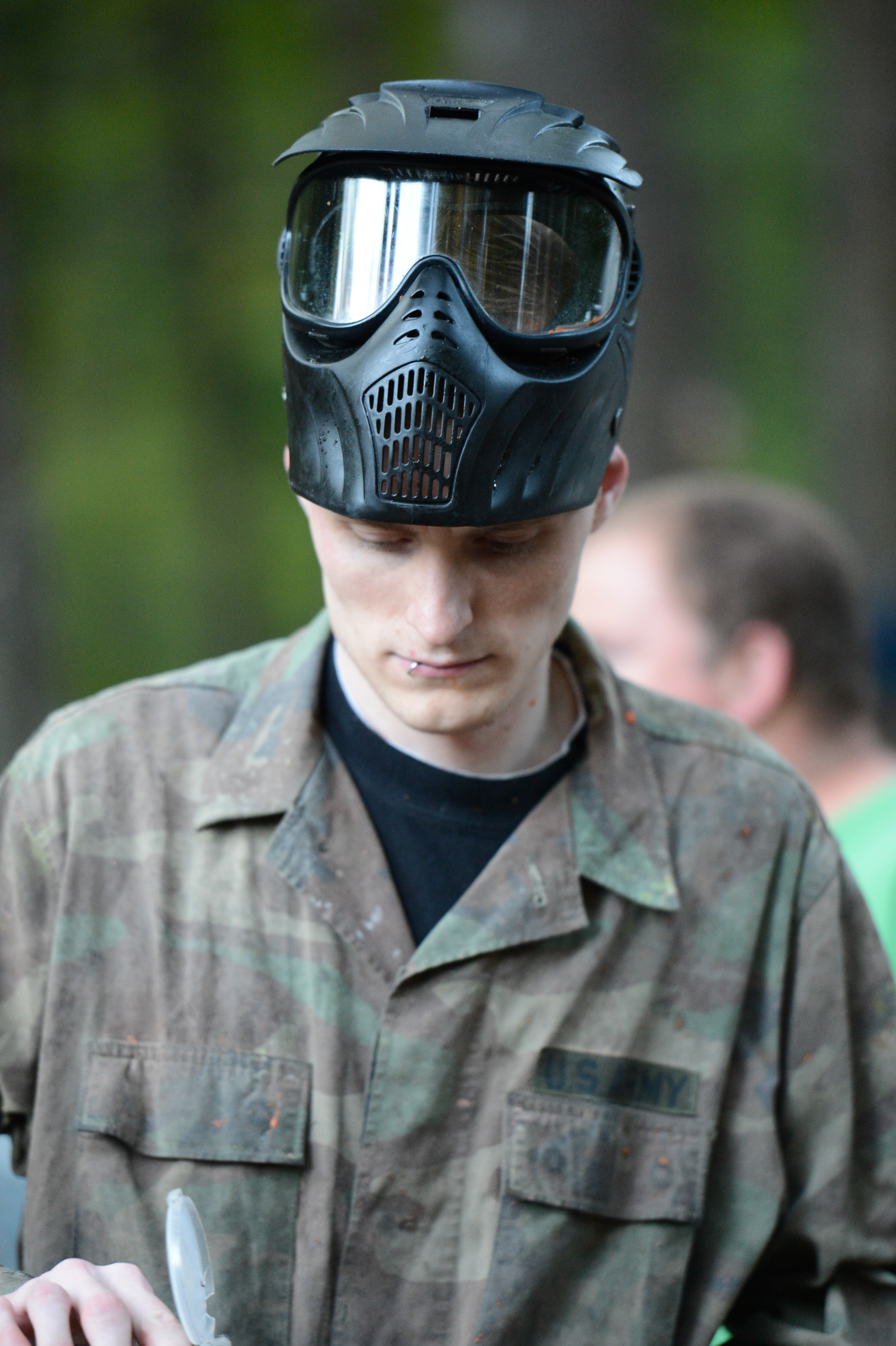 A person wearing a paintball mask and camouflage jacket looks down while handling equipment.