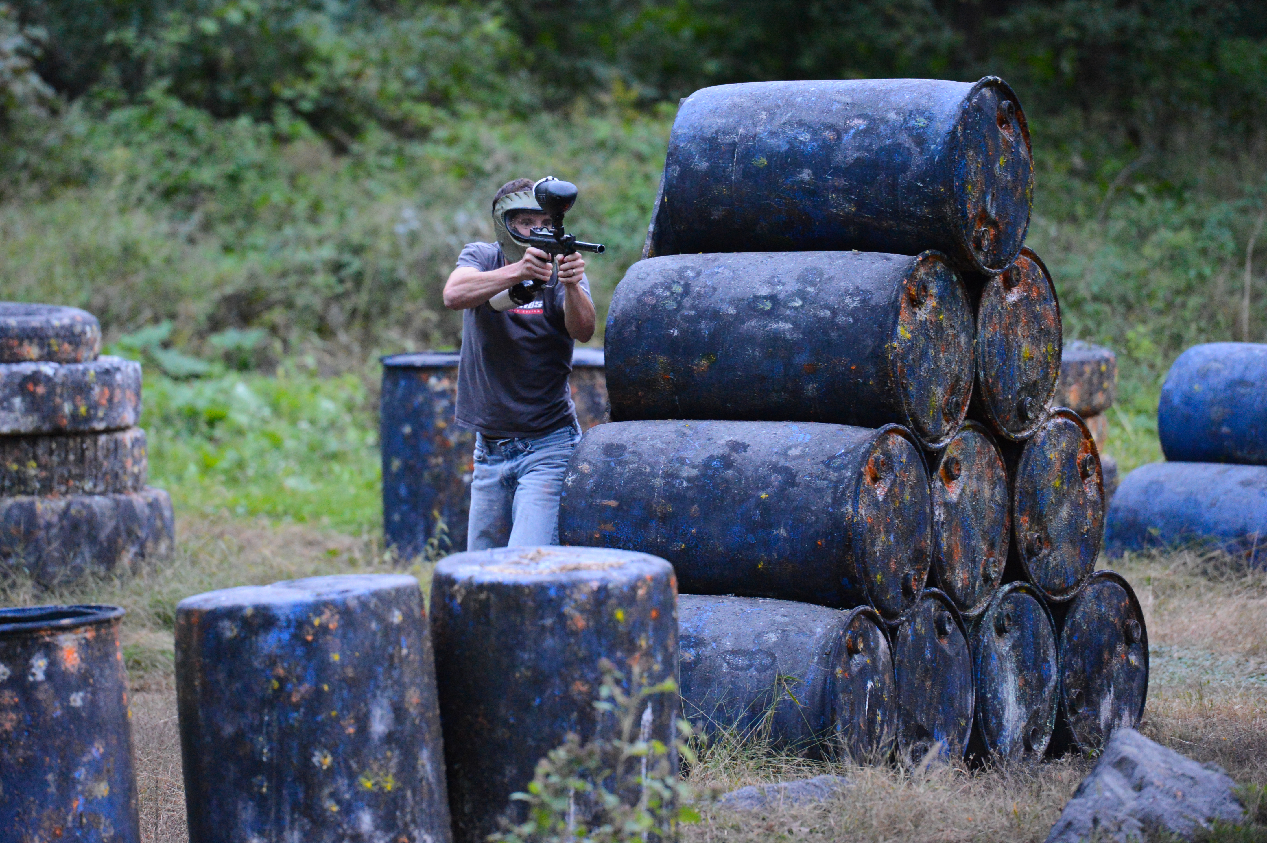 A person wearing a mask aims a paintball gun while taking cover behind stacked barrels in an outdoor field.