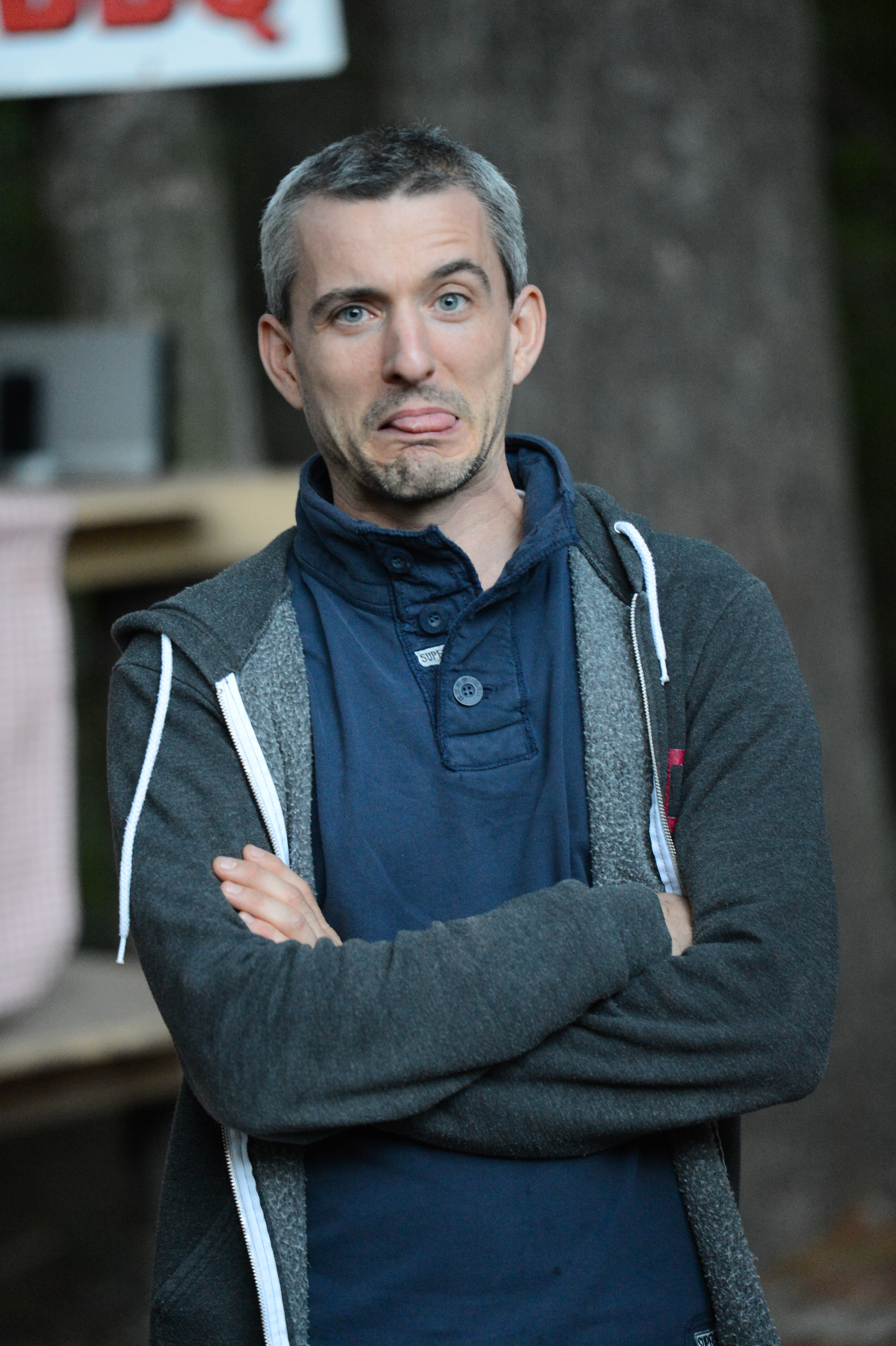 A person with short hair and a hoodie crosses their arms and makes a playful face, sticking out their tongue.