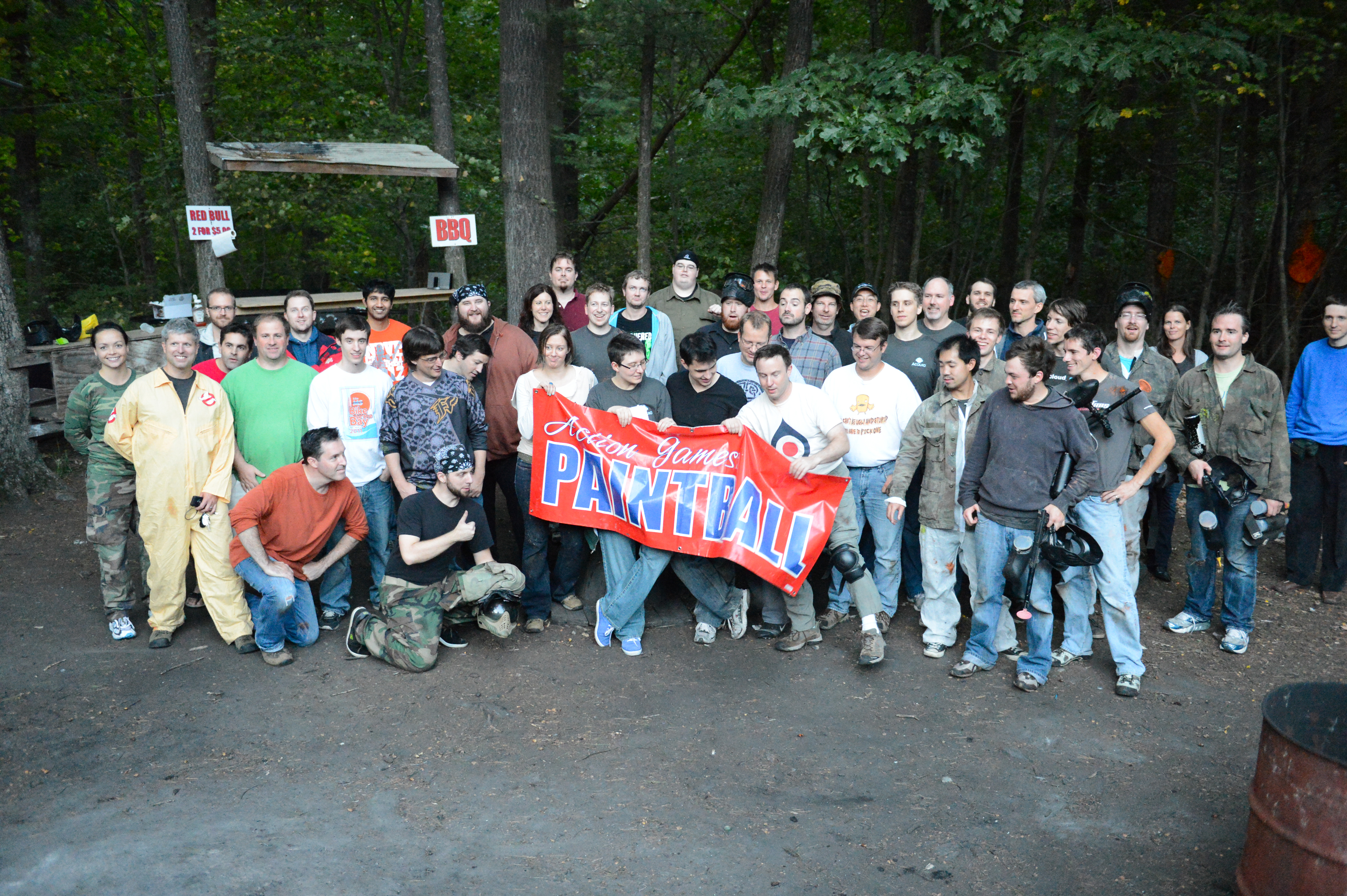 A large group of people poses outdoors, holding a "Paintball" banner, dressed in casual and paintball gear.