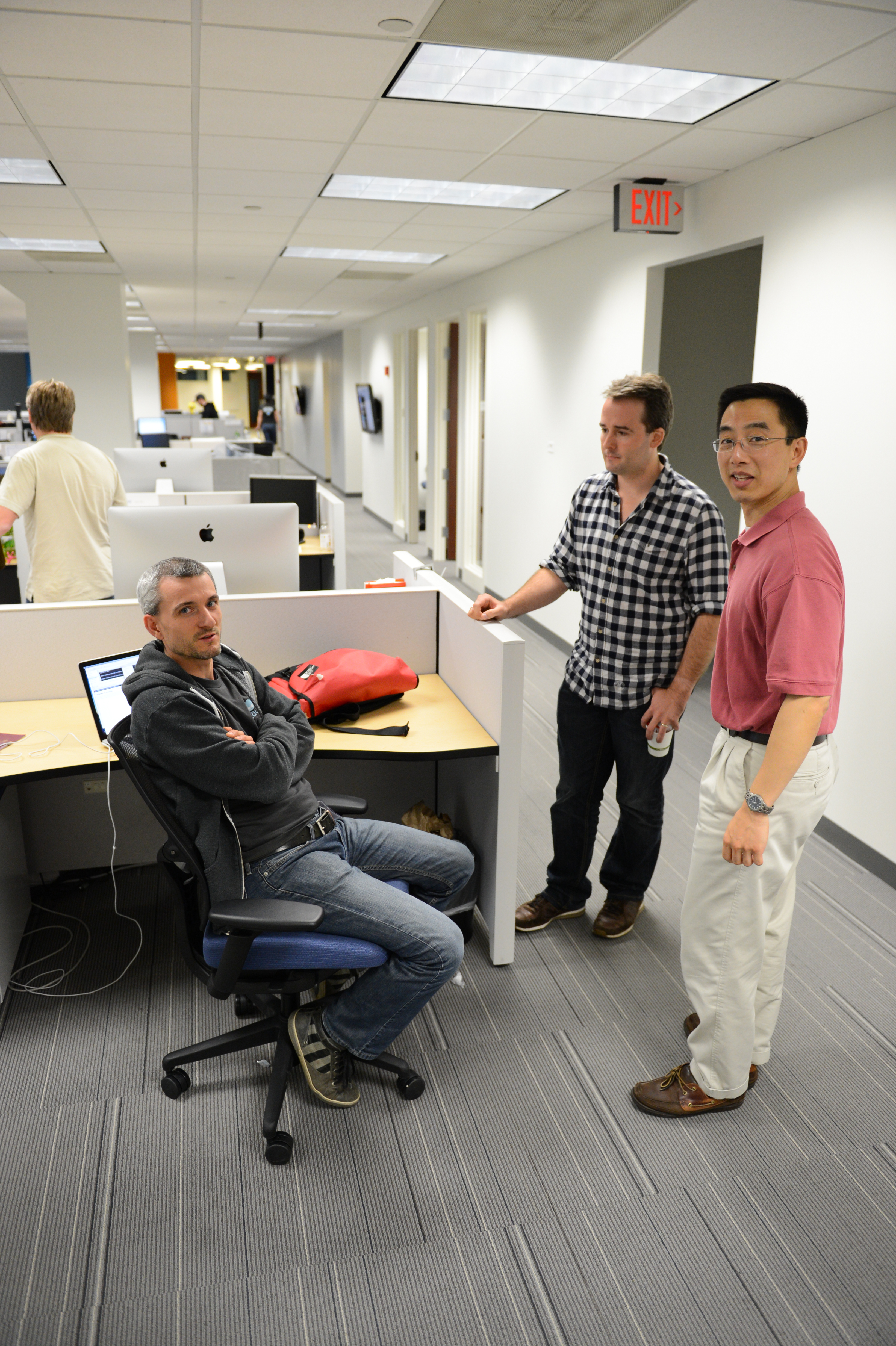 Three men in an office, one sitting at a desk while the other two stand and talk nearby.