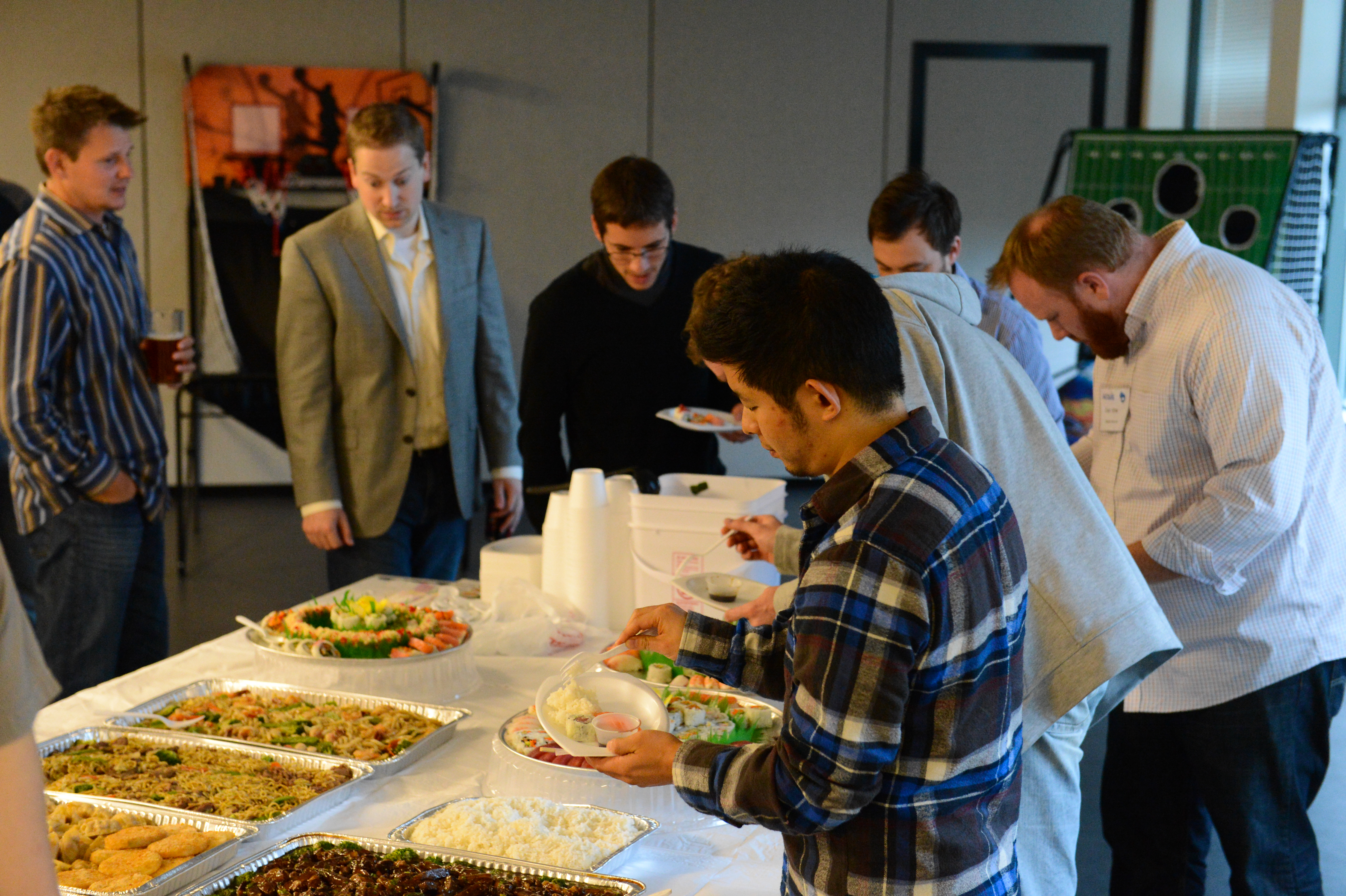 A group of people gathers around a buffet table, serving themselves food at an indoor event.