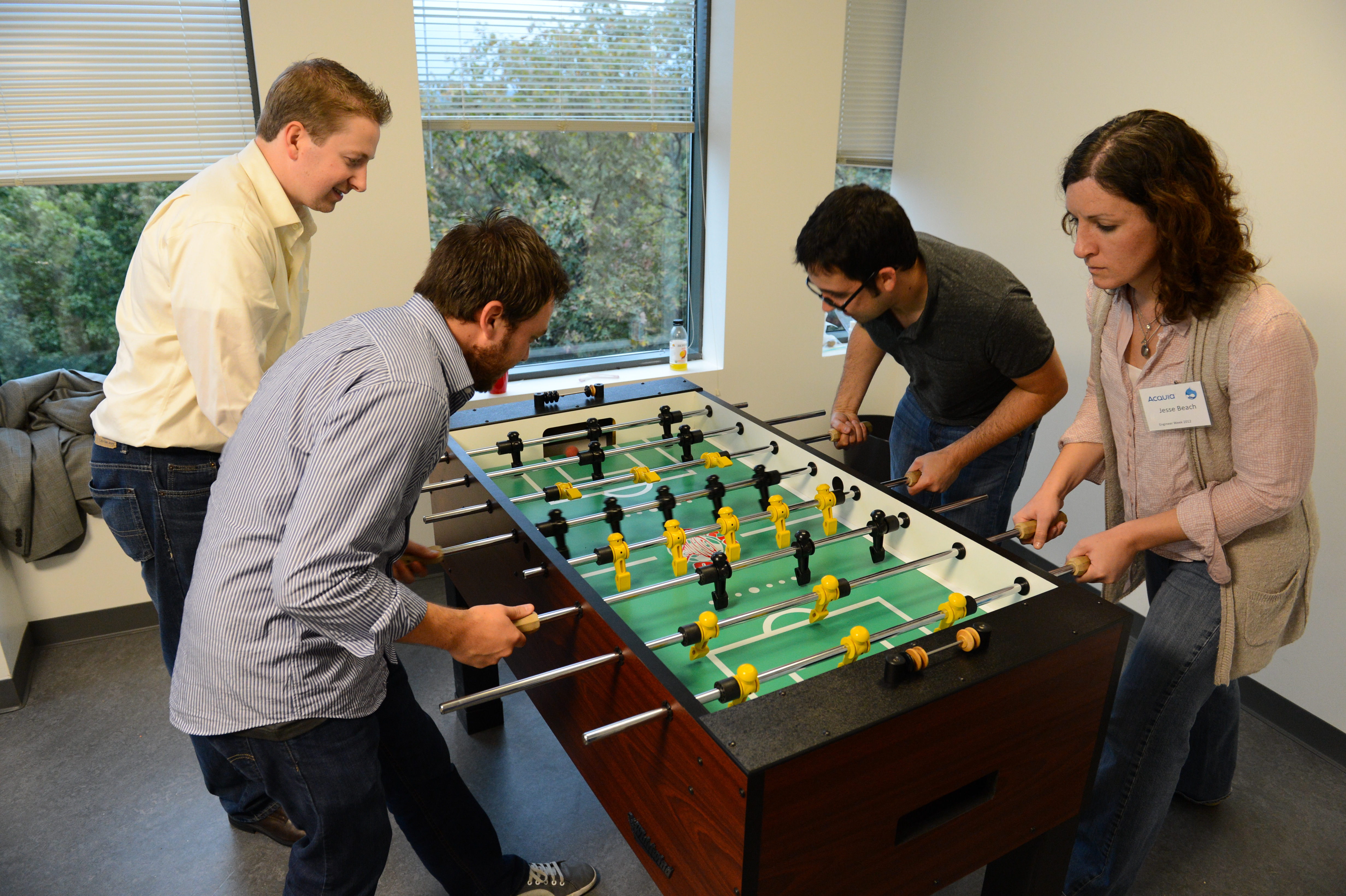 Four people are playing foosball in an office setting, focusing on the game and gripping the handles.