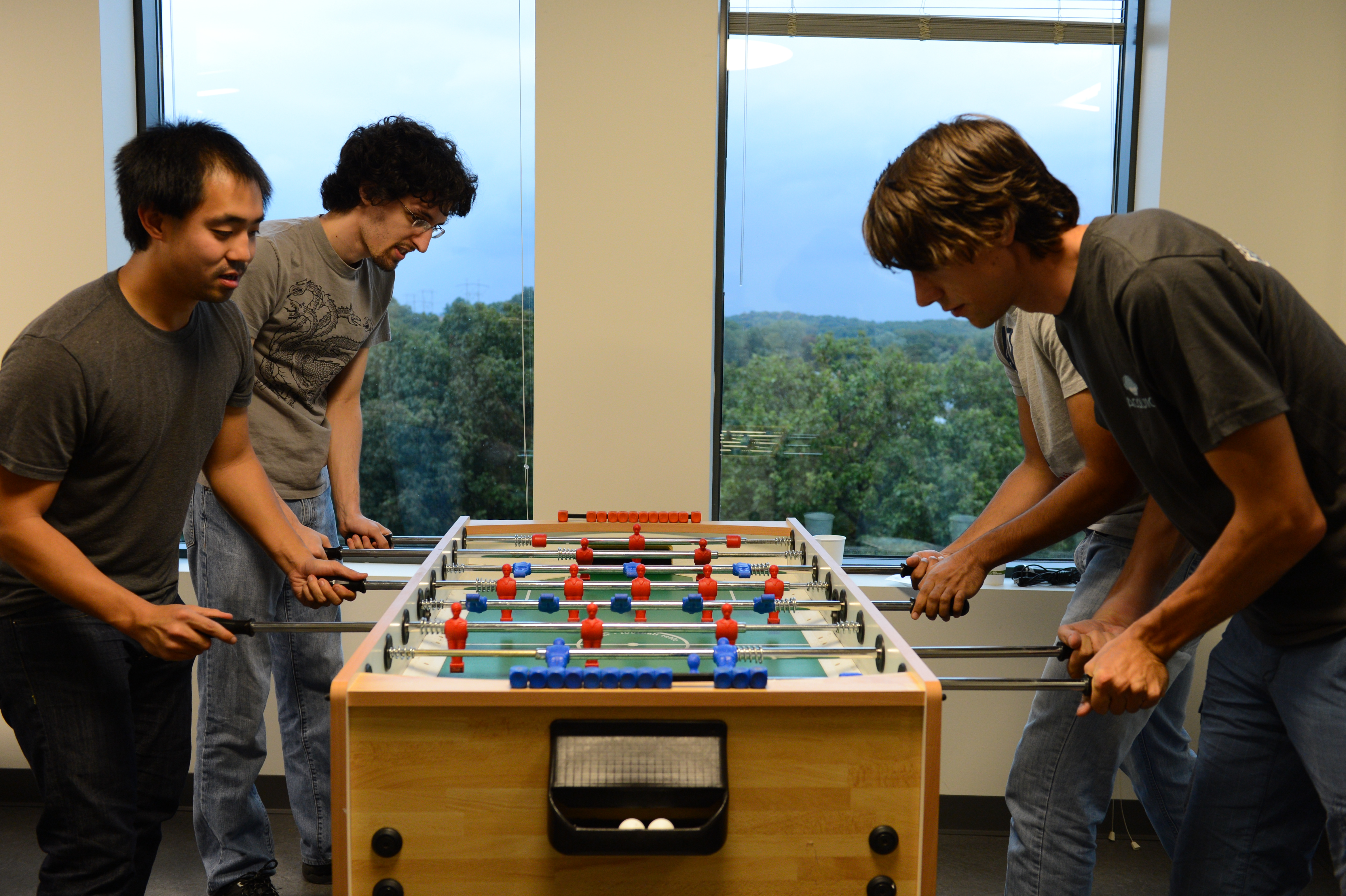 Four people are playing foosball in an office with large windows showing a scenic outdoor view.