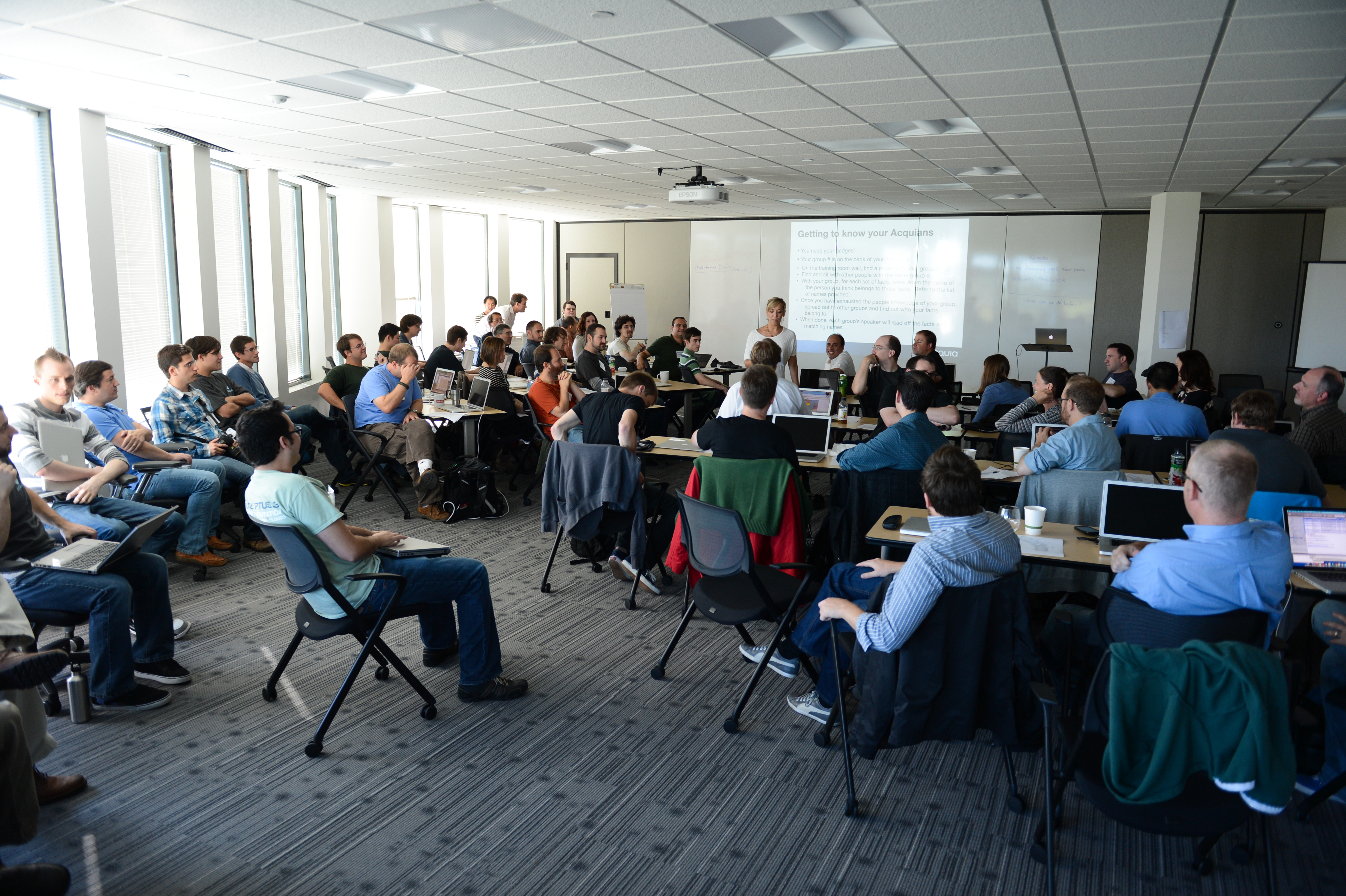 A presenter speaks to a large group of people seated in a conference room, many using laptops.