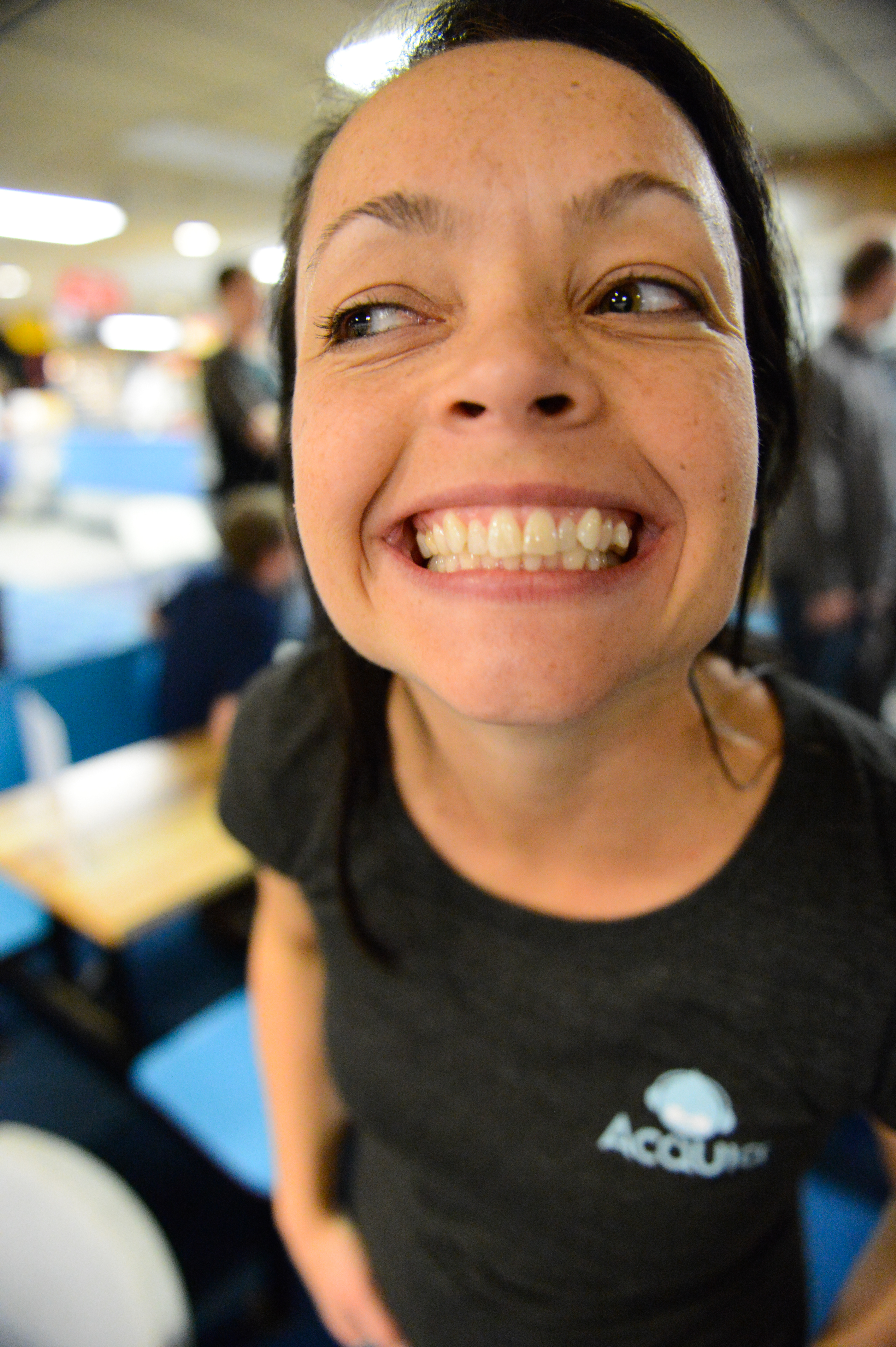 A person wearing an Acquia shirt smiles widely at the camera in a lively indoor setting.