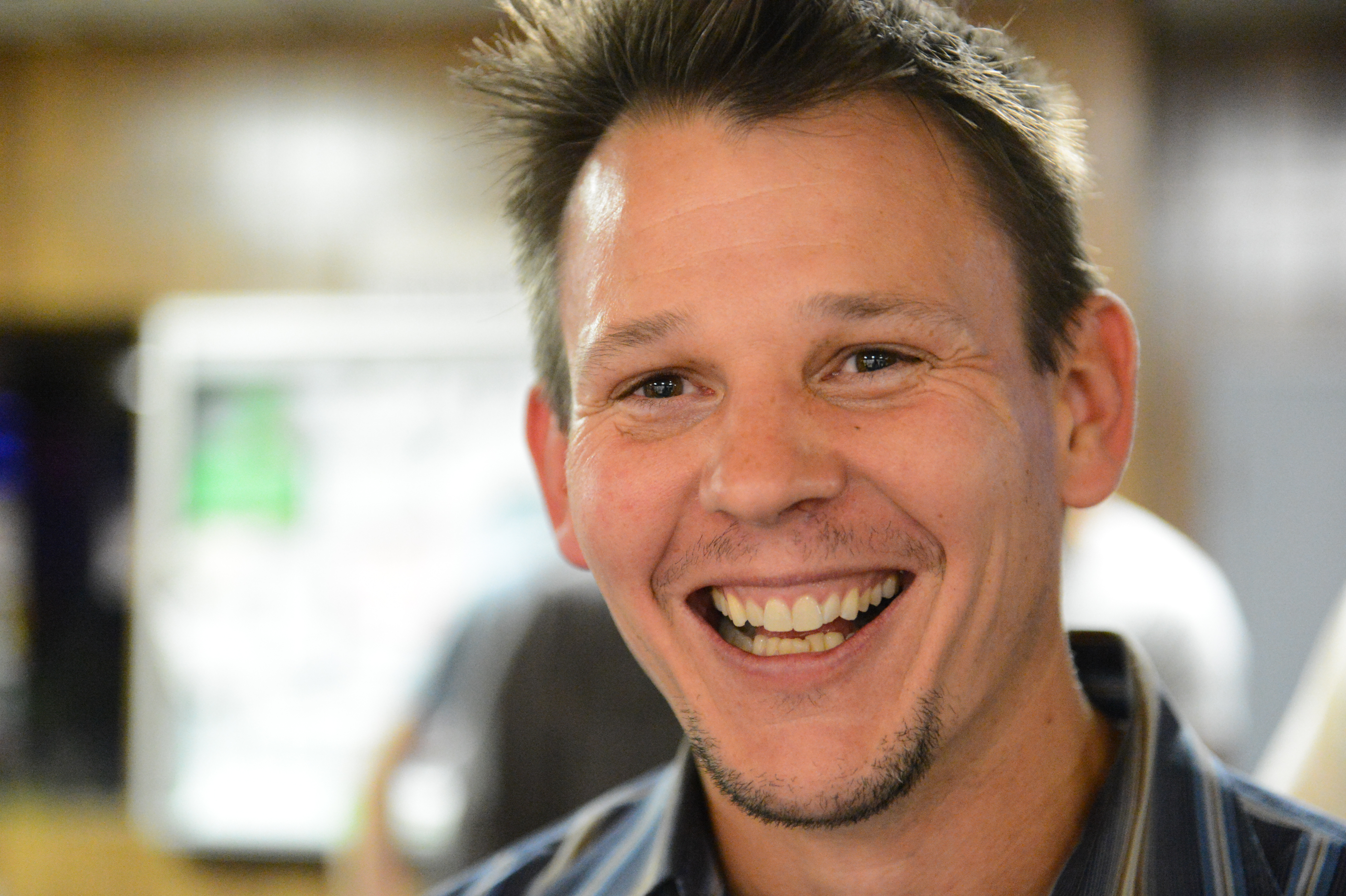 A man with short hair and a striped shirt smiles broadly at the camera in an indoor setting.