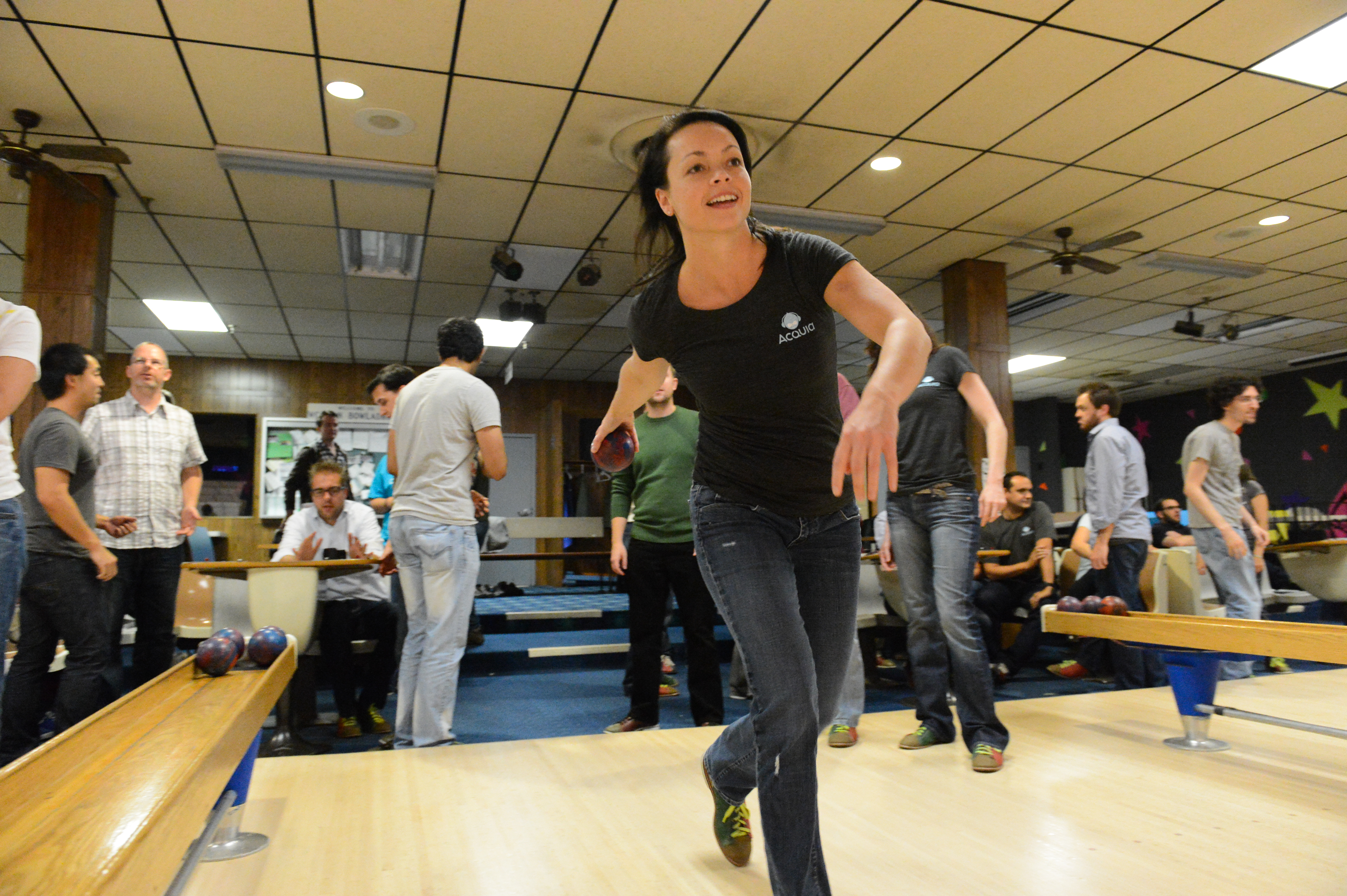 A woman in a black Acquia shirt releases a bowling ball, surrounded by colleagues at a bowling alley.