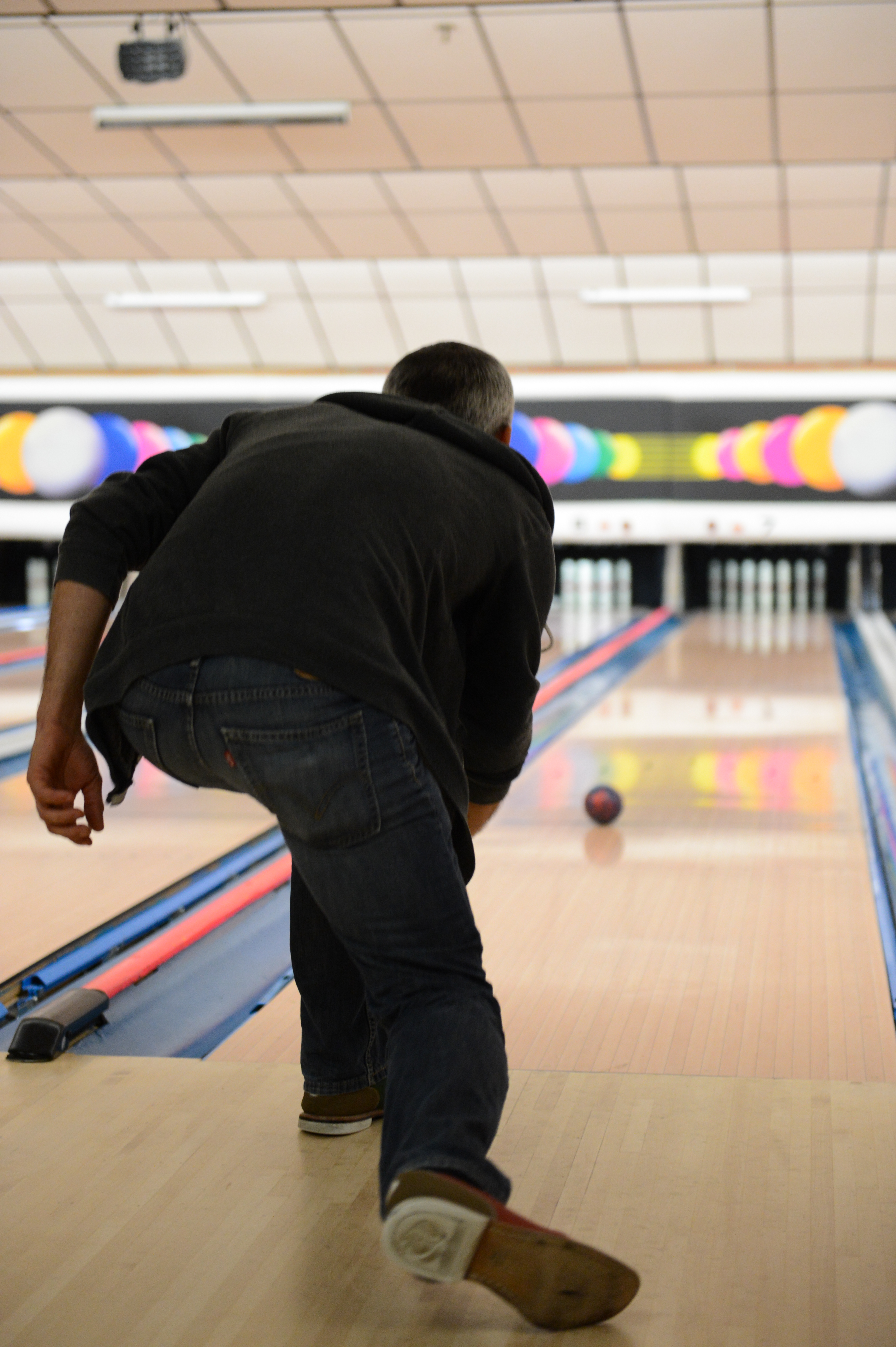 A person in a dark jacket and jeans bowls a ball down a lane at a bowling alley.