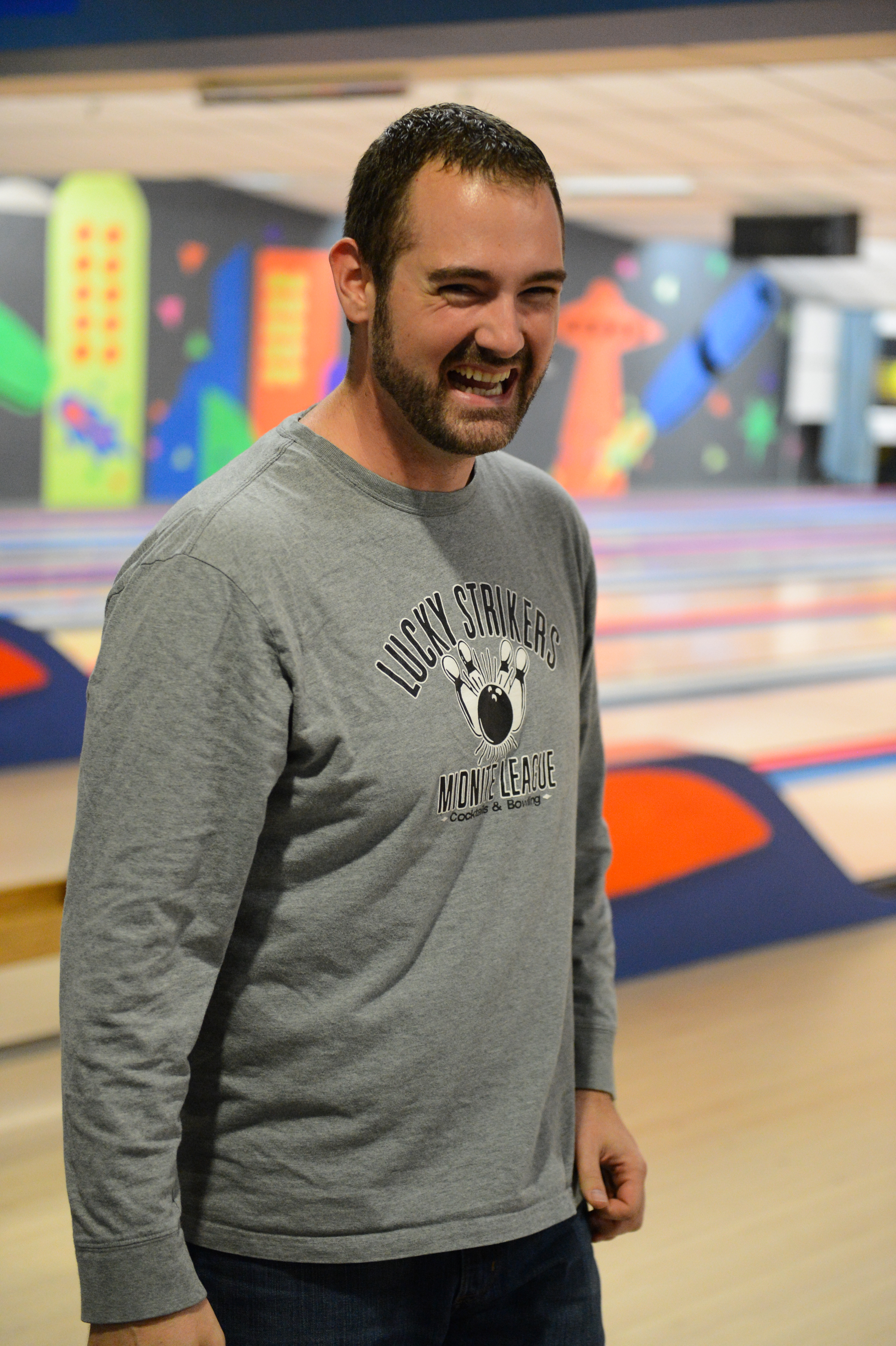 A man in a gray bowling league shirt smiles while standing in a bowling alley.