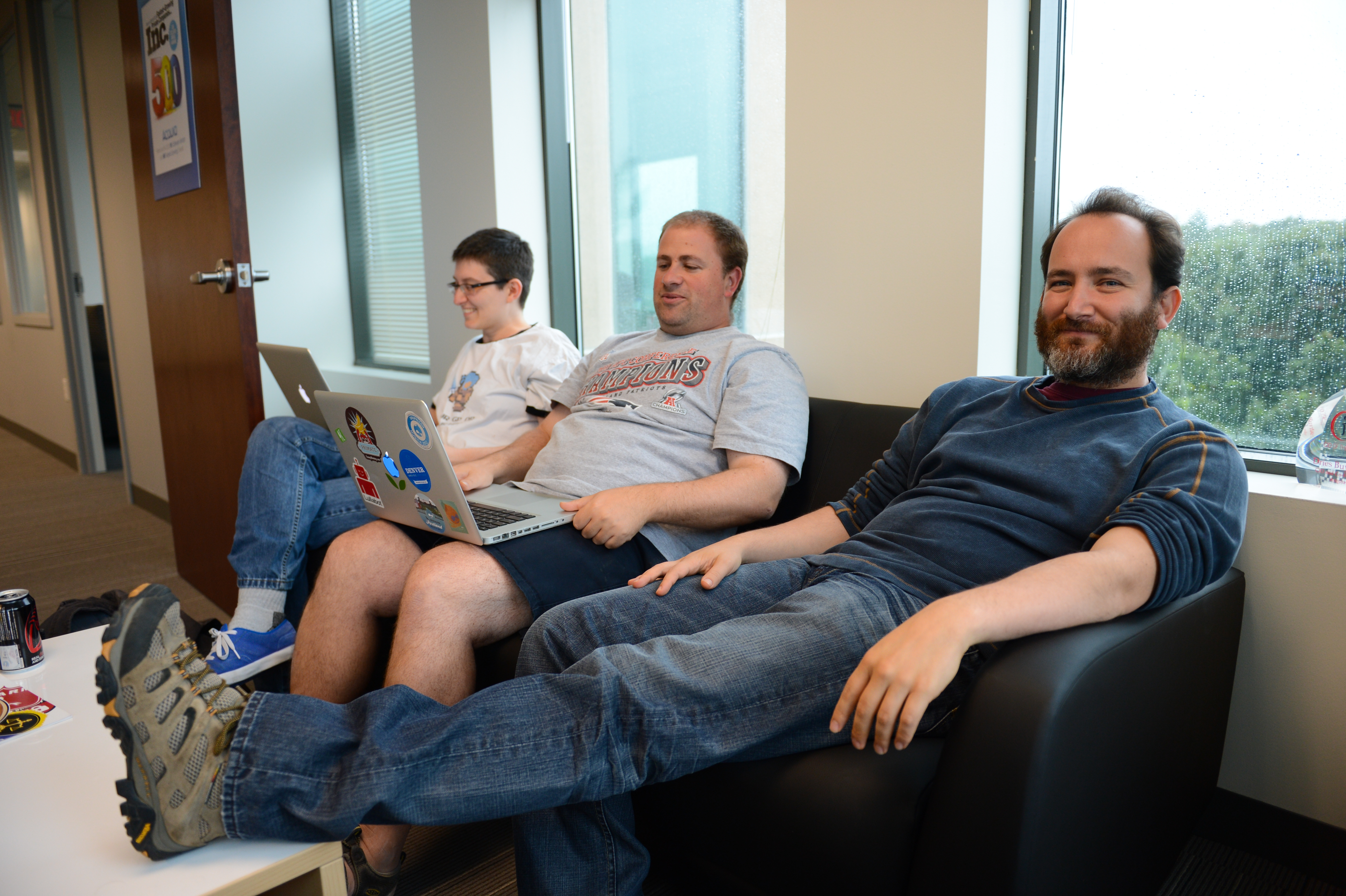 Three people sitting in an office lounge, two using laptops while one relaxes with feet on a table.