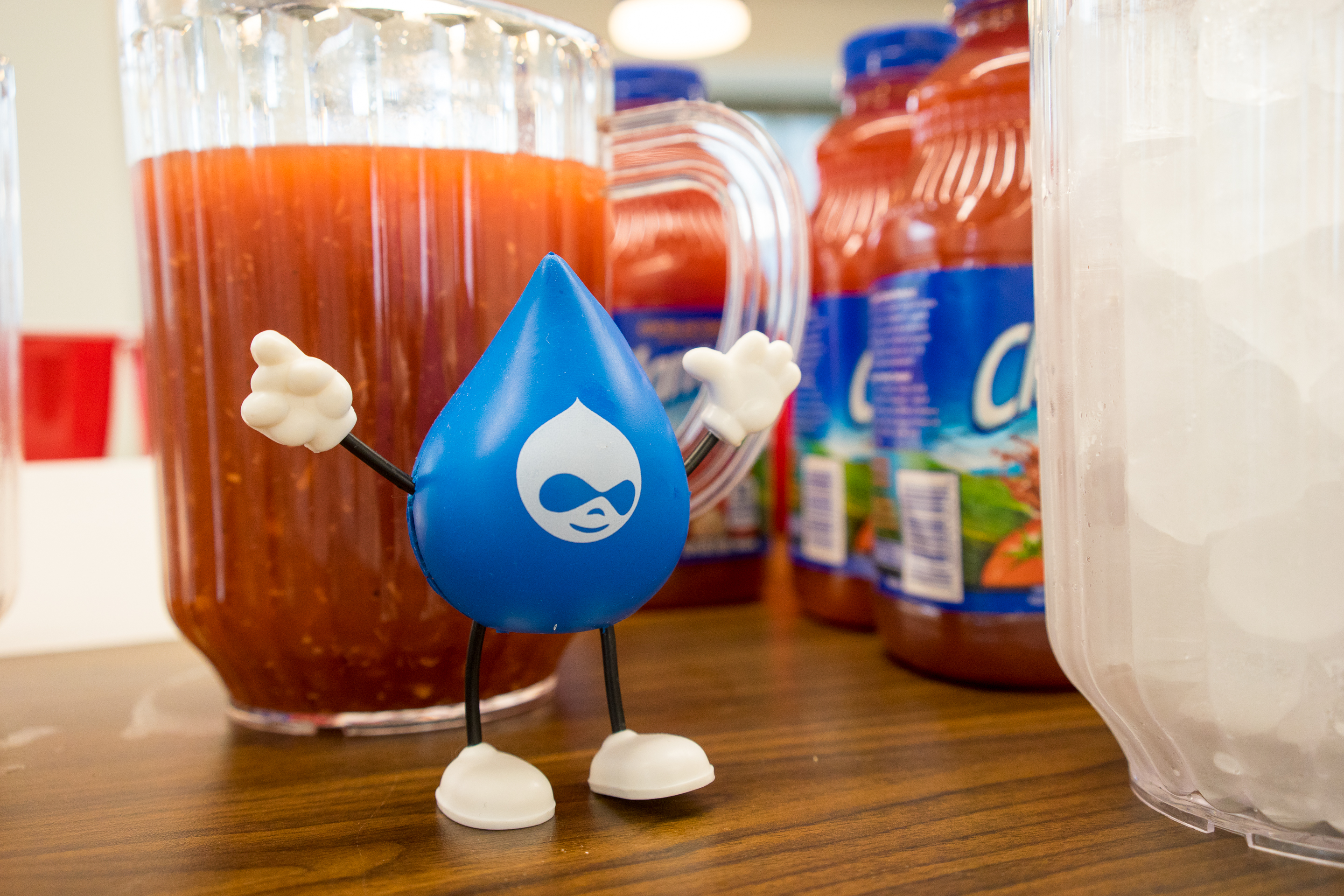 A small blue Acquia mascot figurine stands on a table near pitchers of juice and bottles of Clamato.