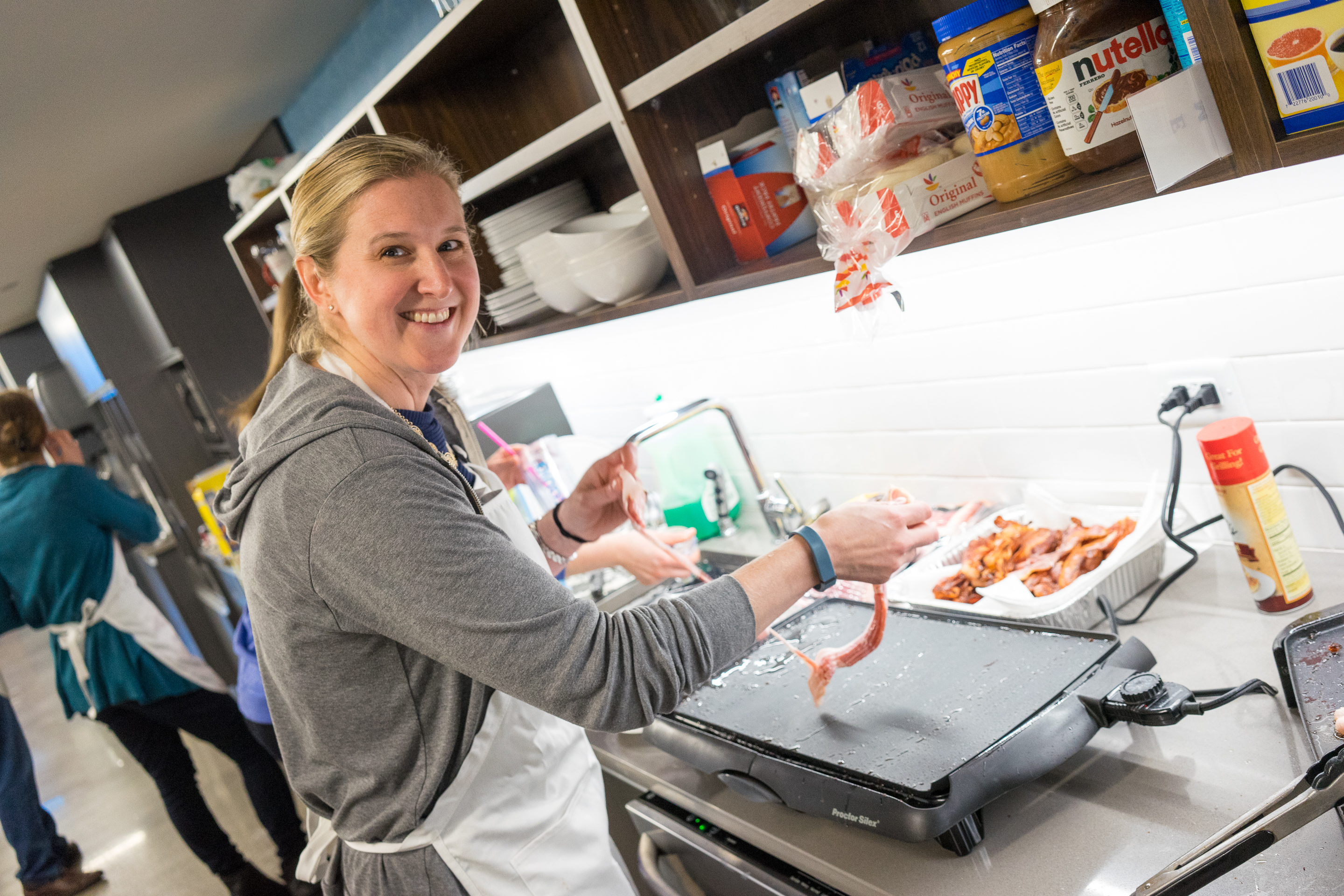 A woman wearing an apron smiles while cooking bacon on a griddle in a kitchen.