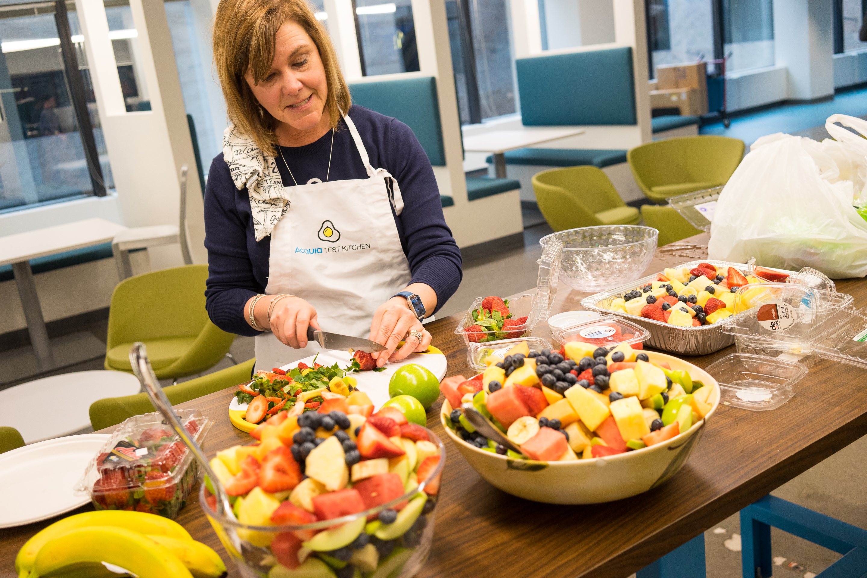 A woman wearing an "Acquia Test Kitchen" apron prepares fresh fruit salad at a table with various ingredients.