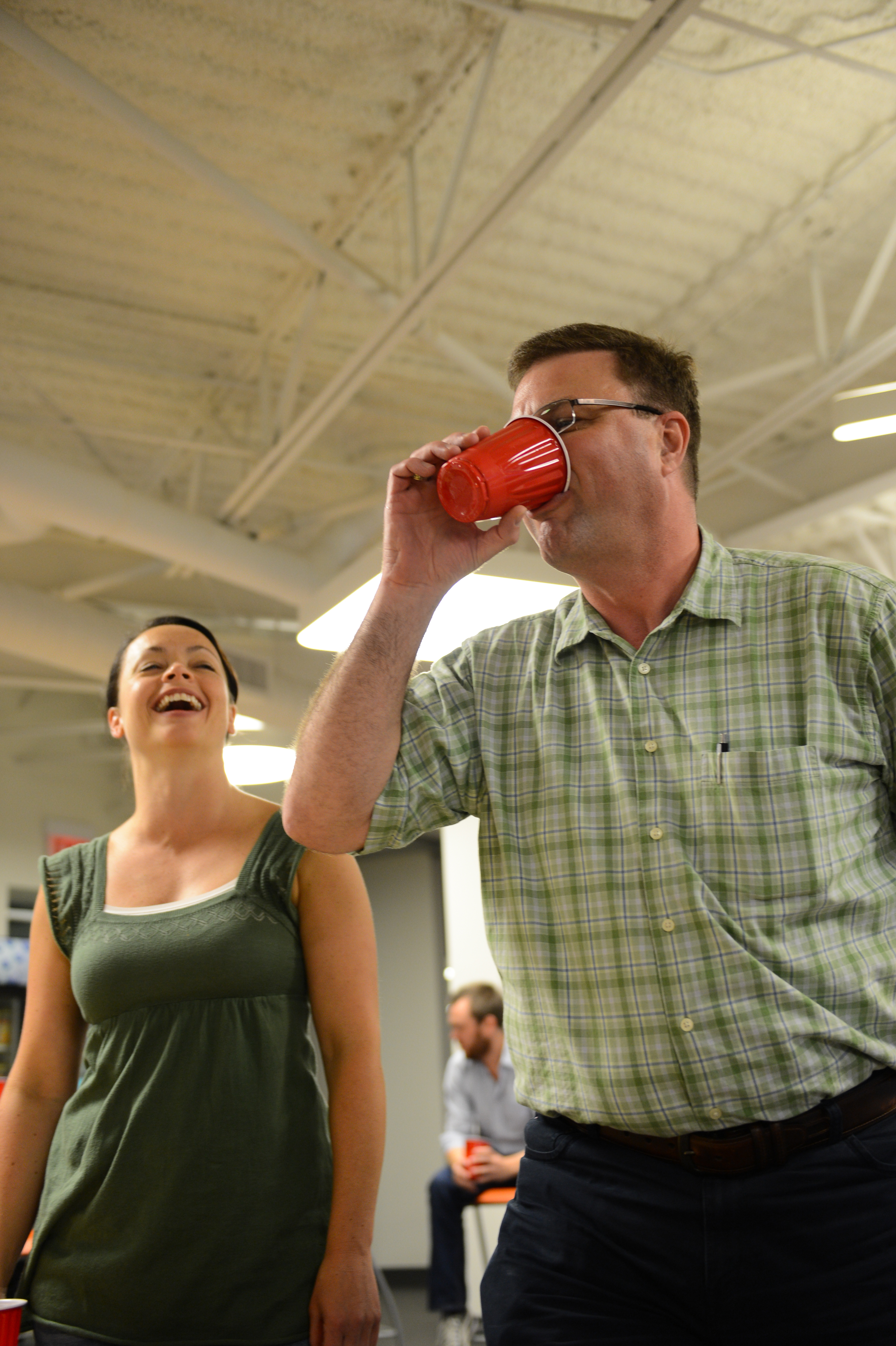 A man in a plaid shirt drinks from a red cup while a woman nearby laughs.