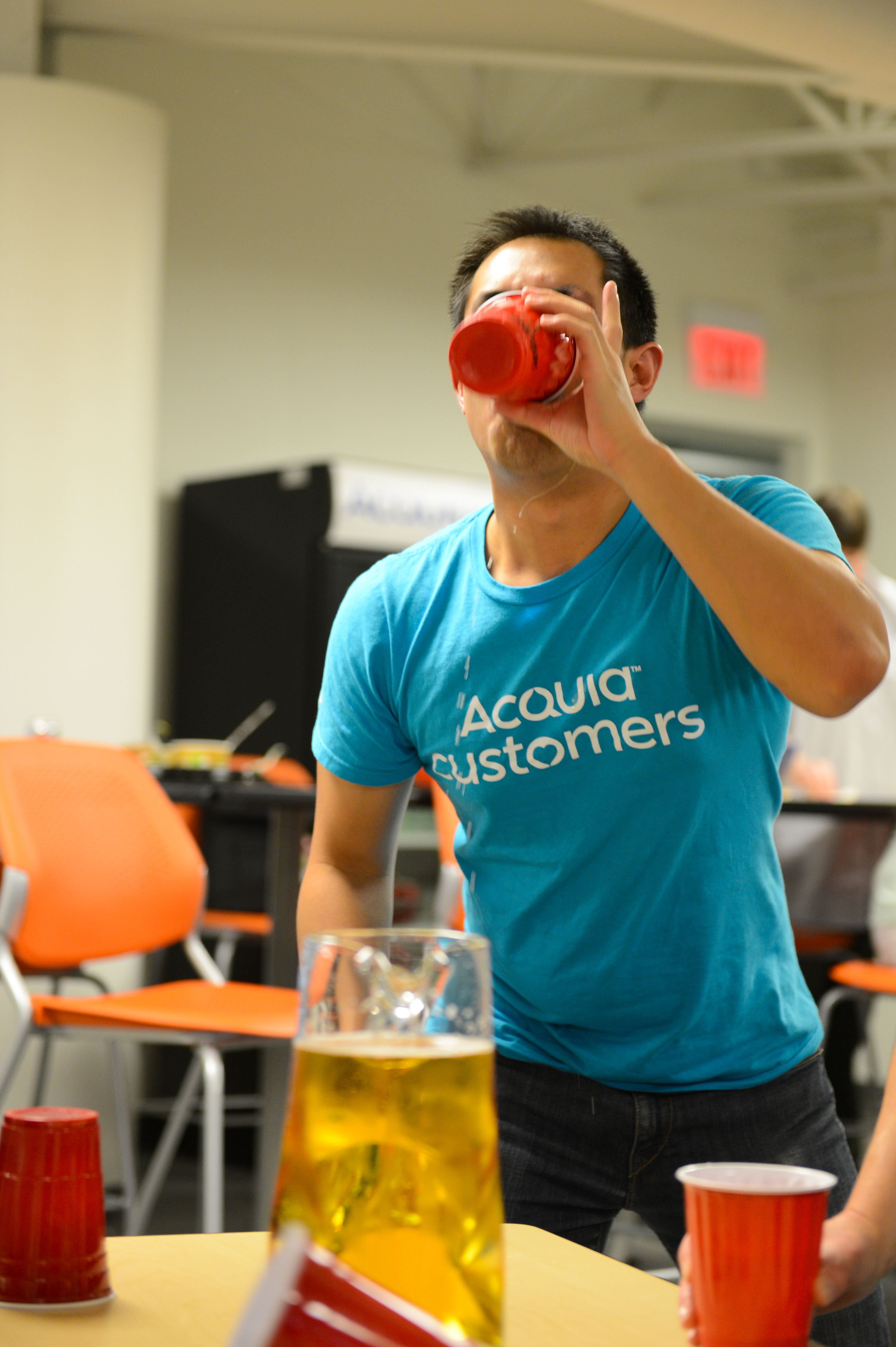 A person in a blue shirt drinks from a red cup at a party with drinks on the table.