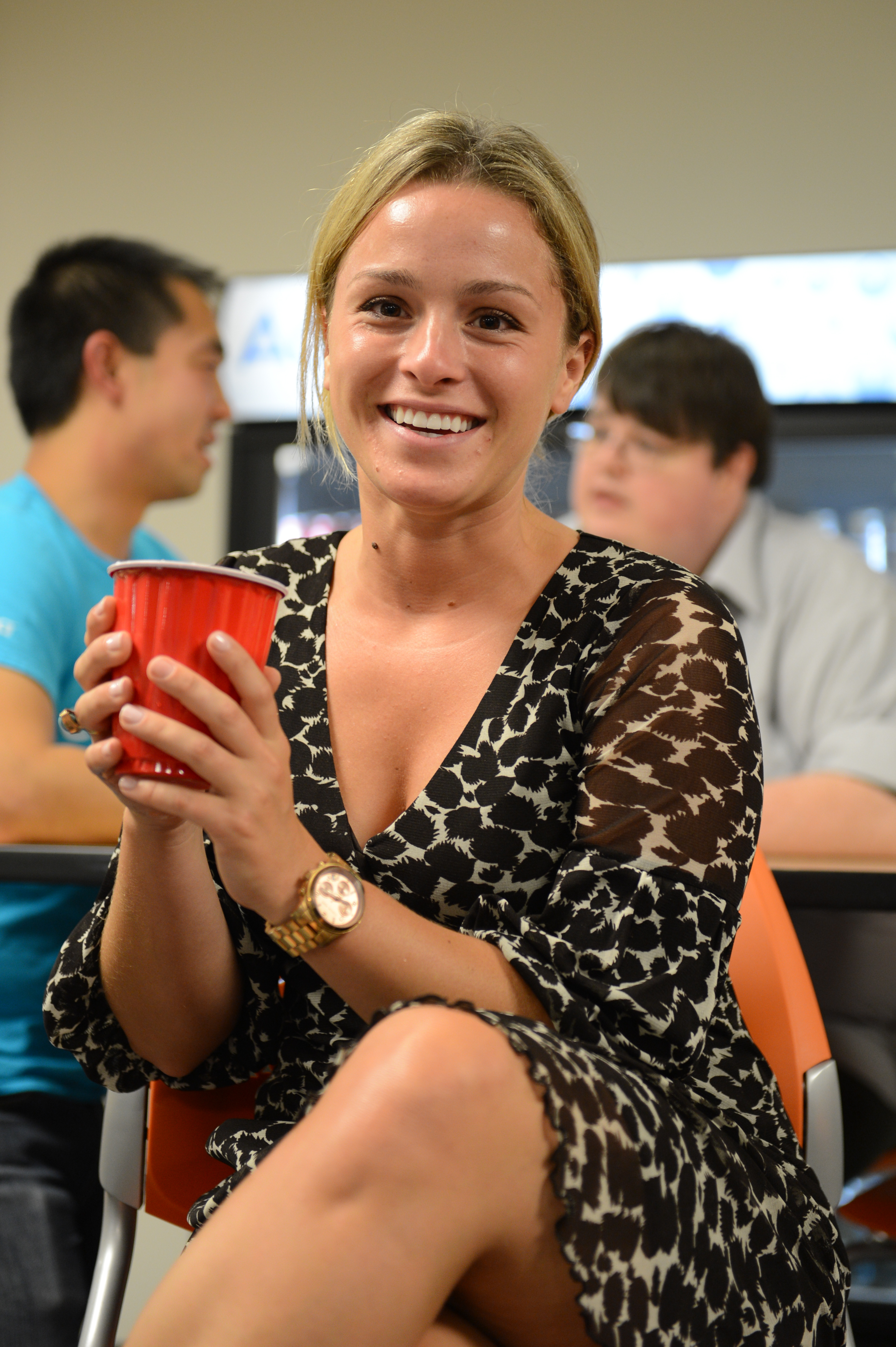 A smiling woman holds a red cup at a party, with people socializing in the background.