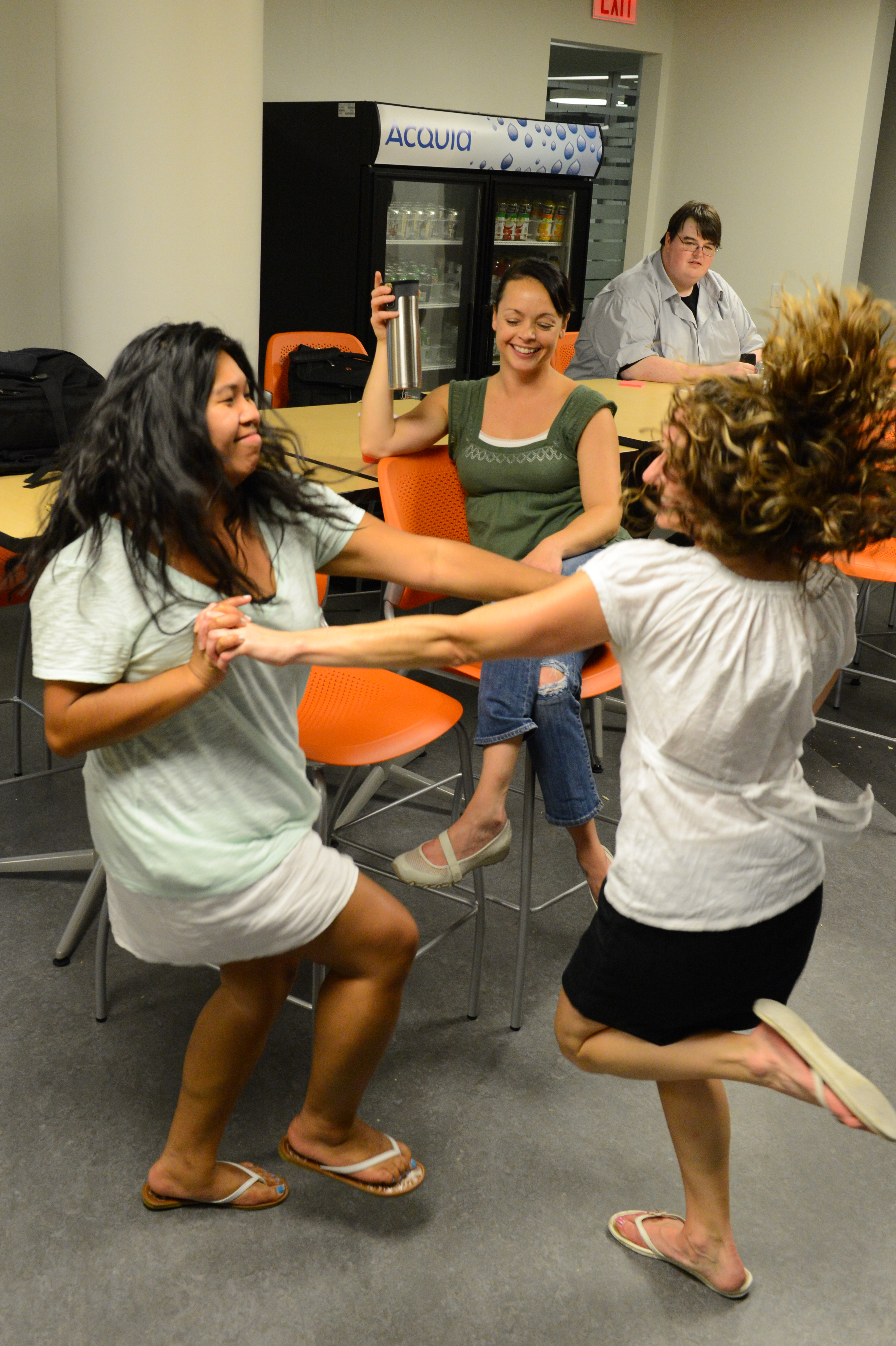 Two women dance energetically while holding hands, as another person watches and smiles in a casual indoor setting.