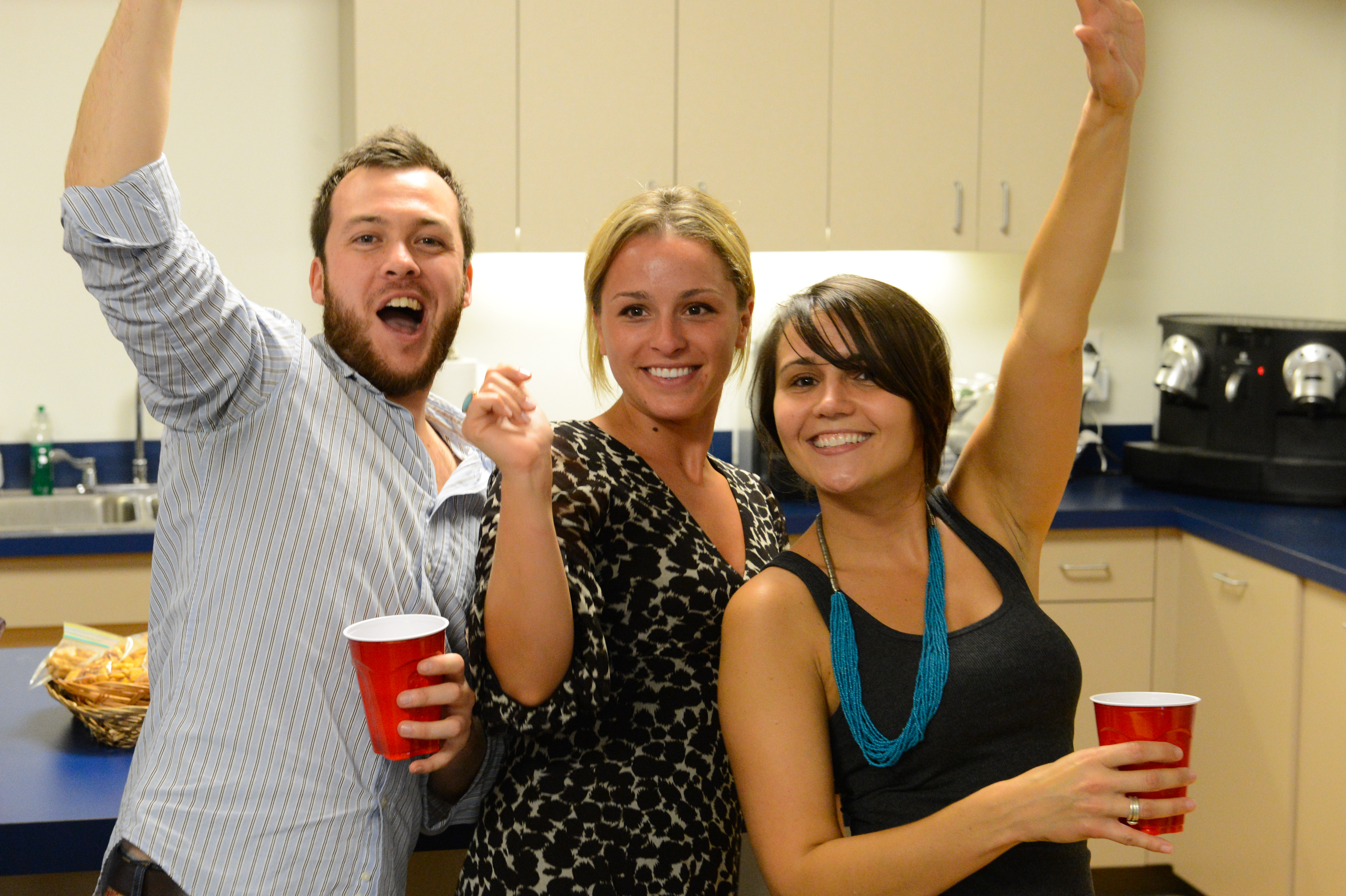 Three people smiling and raising their arms while holding red cups at a party in a kitchen.