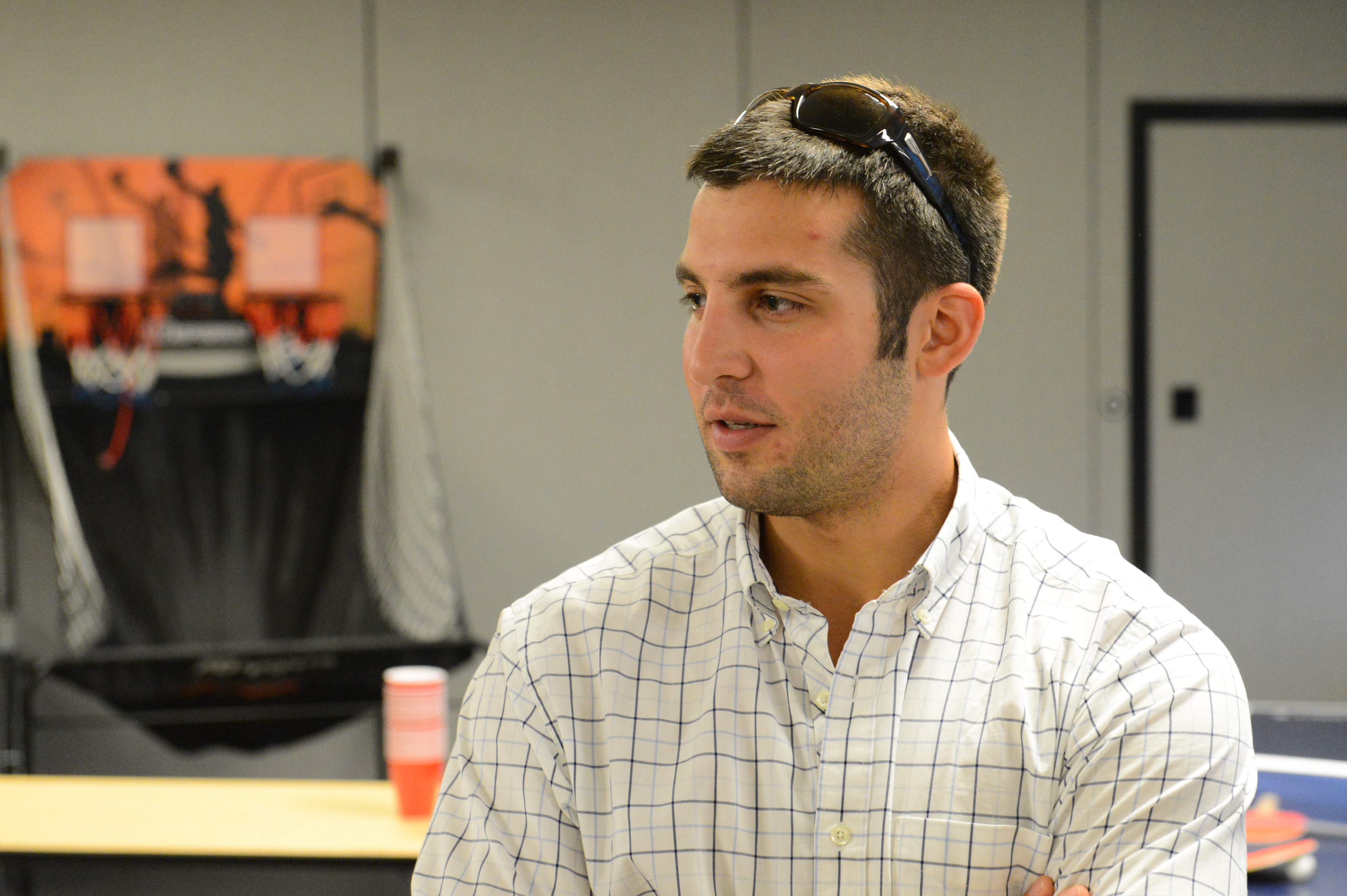 A man in a checkered shirt with sunglasses on his head stands and listens at a party.