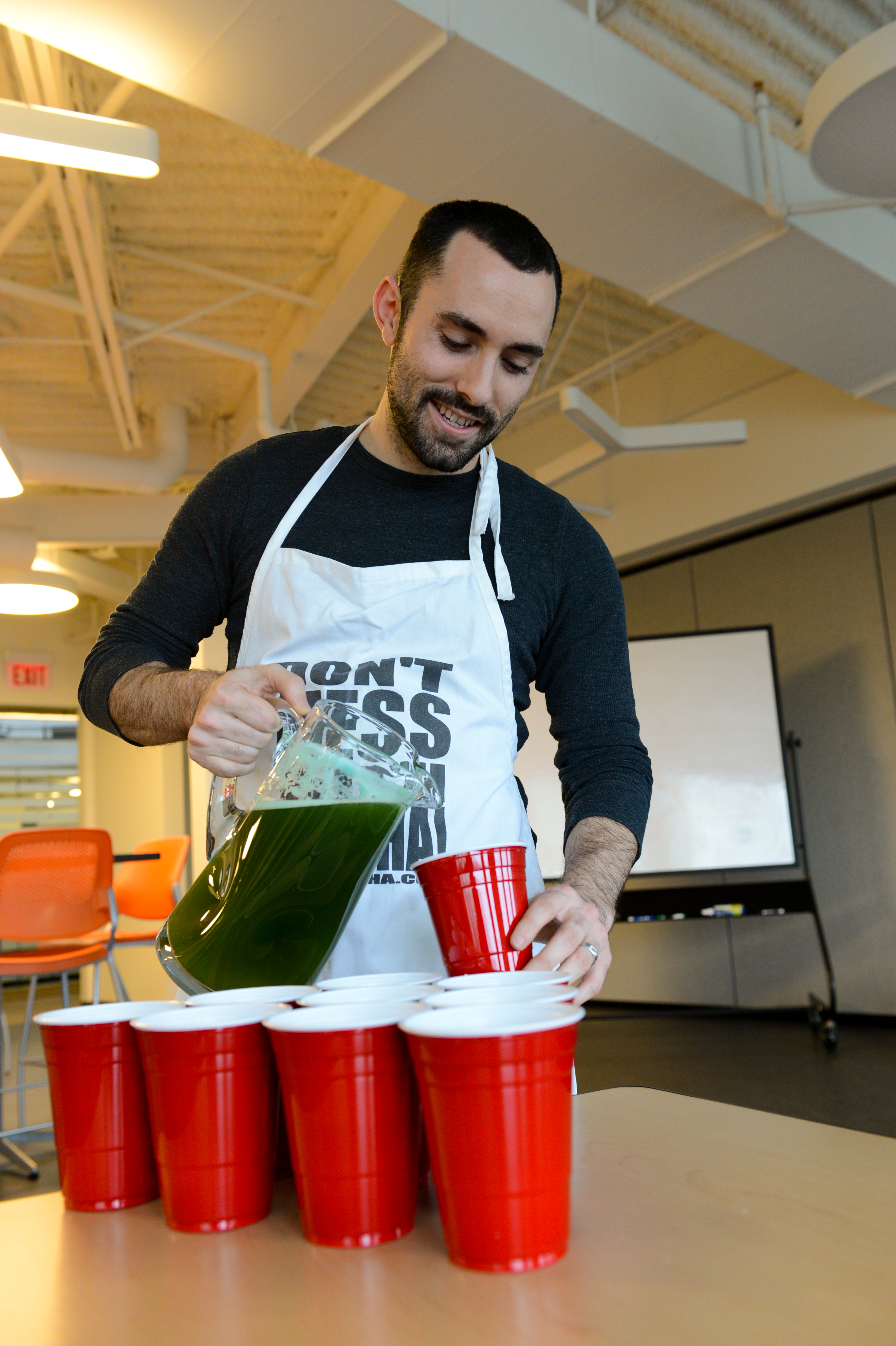 A man wearing an apron pours a green drink from a pitcher into red plastic cups at a party.