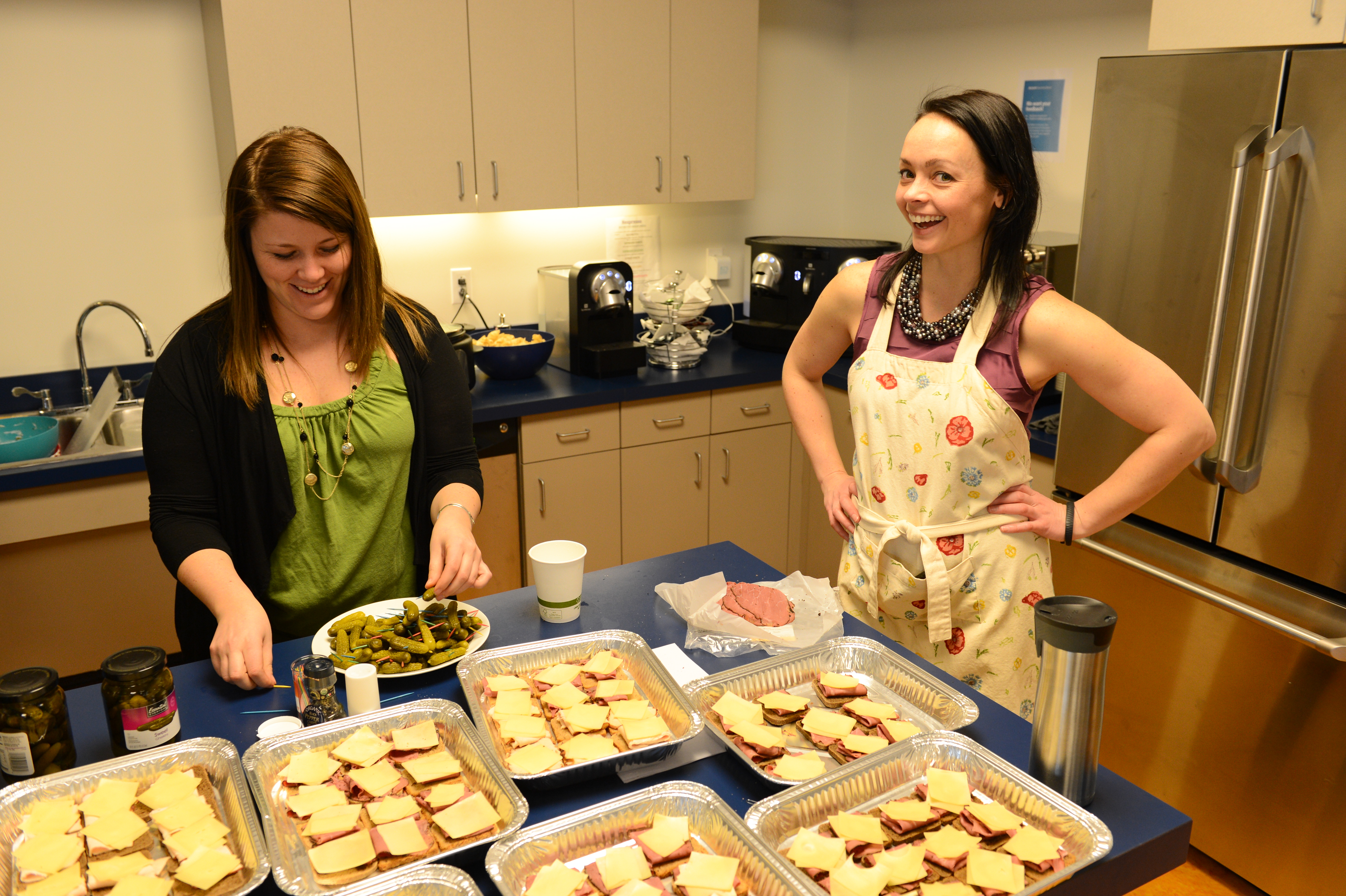 Two women in a kitchen preparing food, one wearing an apron and smiling while the other arranges pickles on a plate.