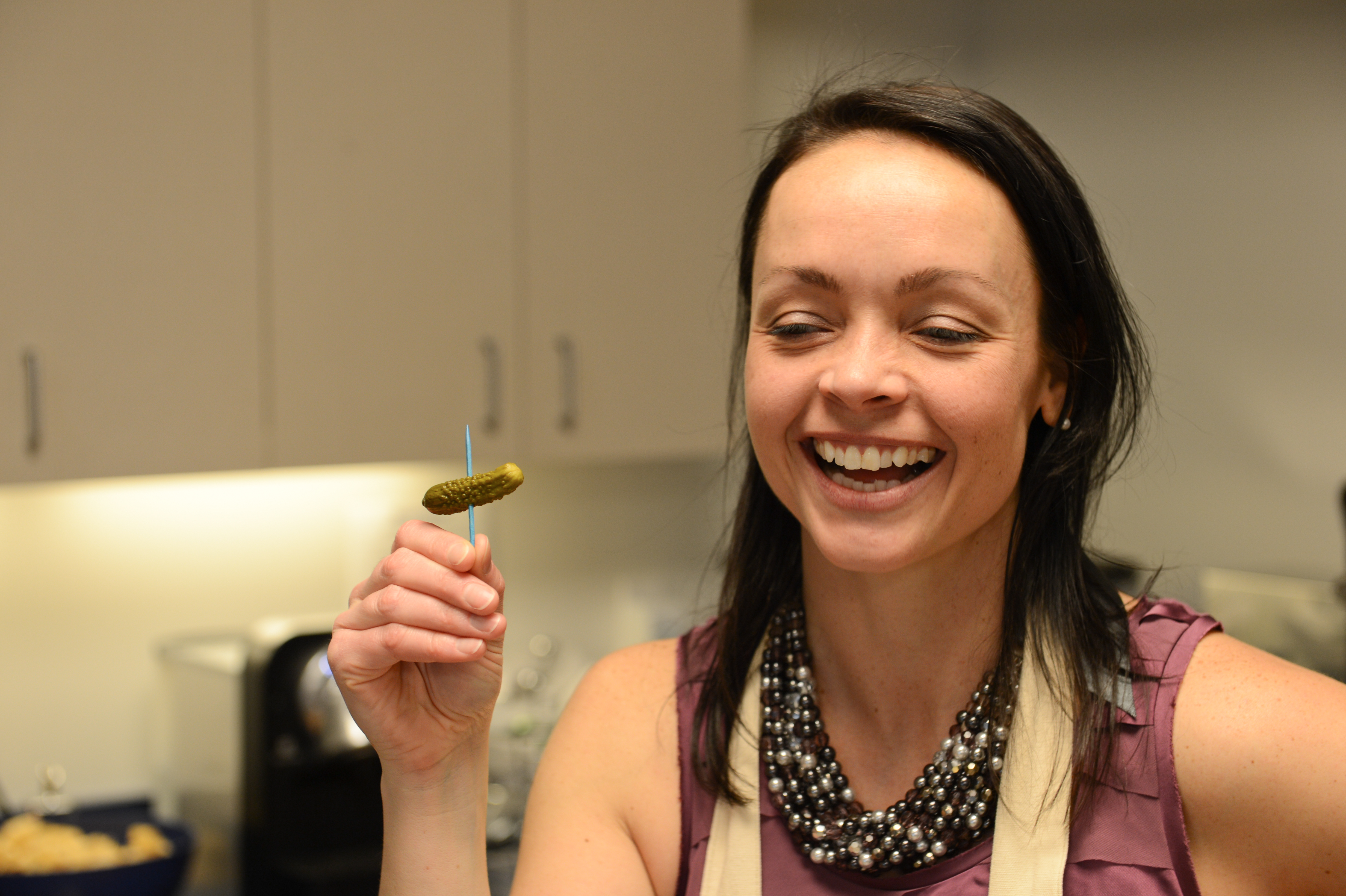 A smiling woman holds a small pickle on a toothpick at a party in a kitchen setting.