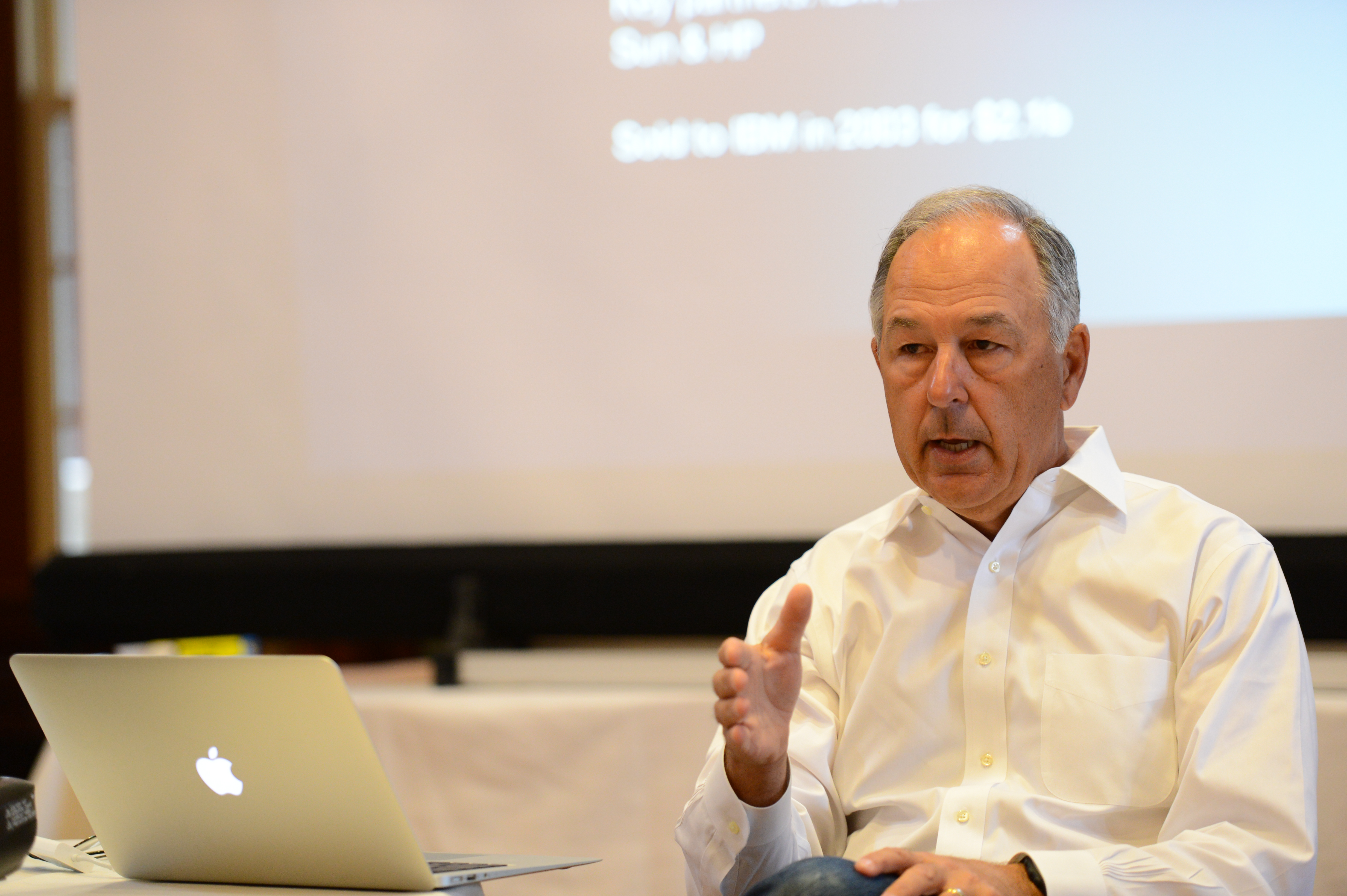 A man in a white shirt speaks while gesturing, with a laptop and presentation screen in the background.