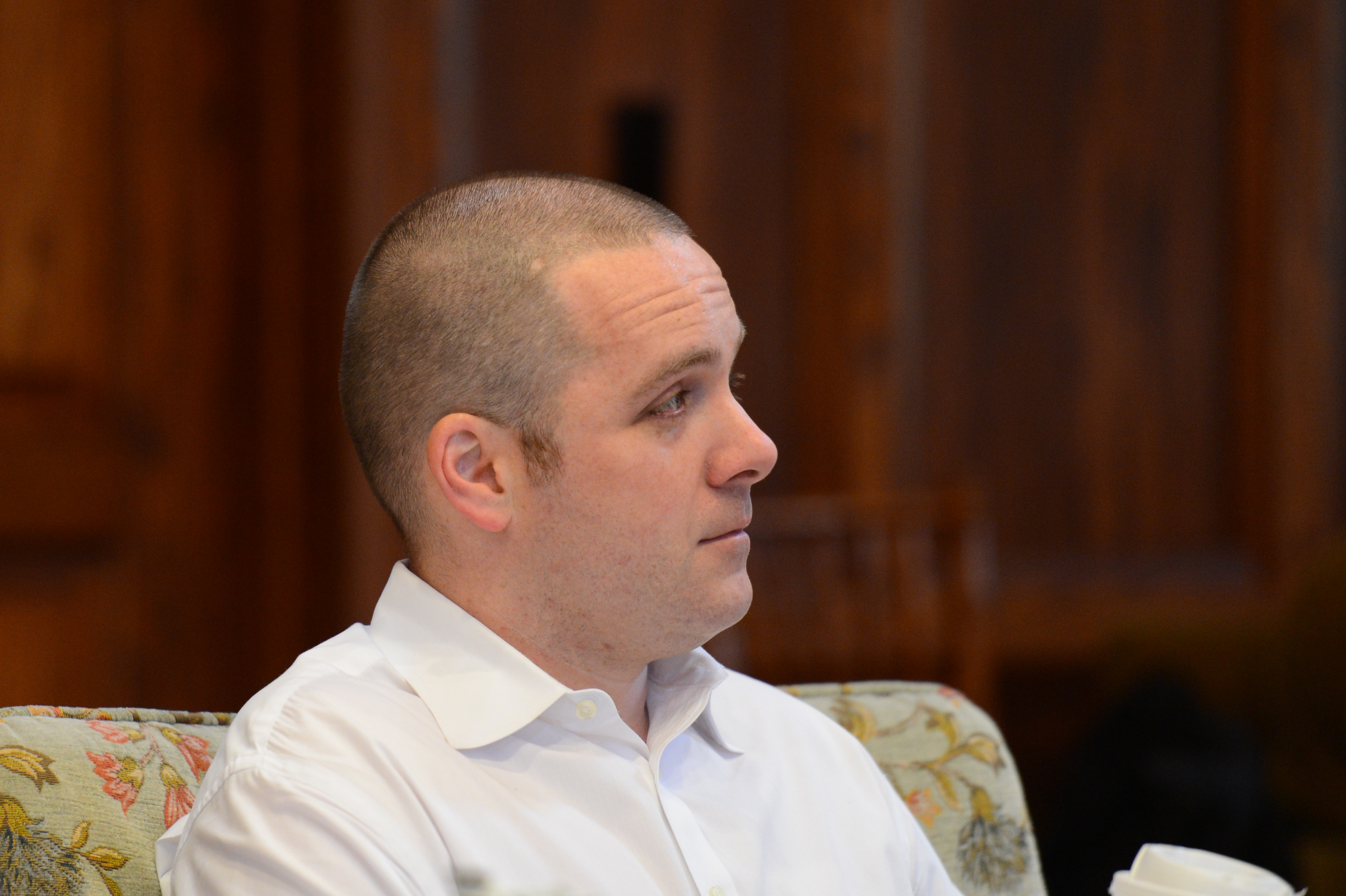 A man in a white shirt sits and listens attentively, holding a cup in his hand.