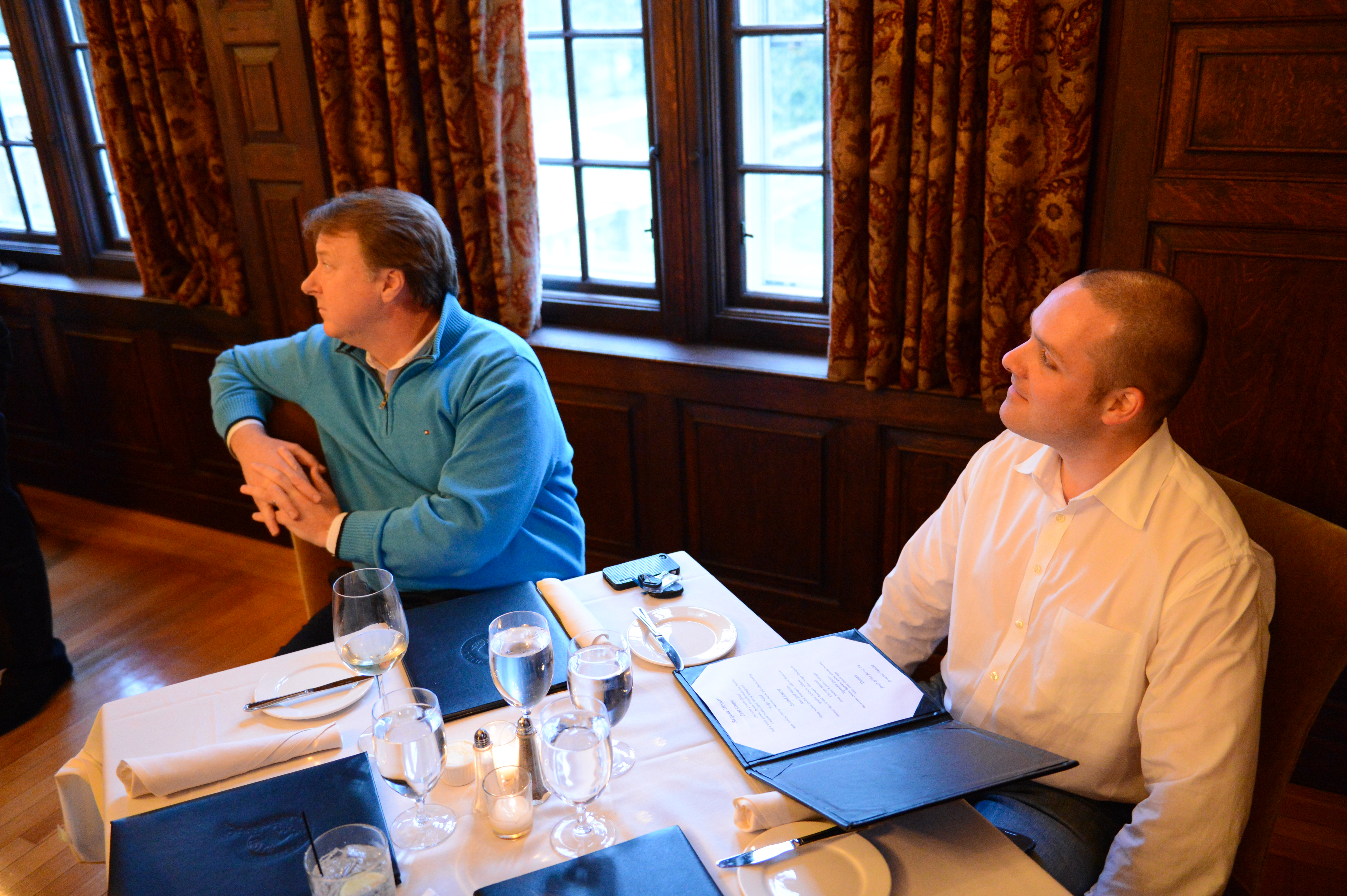 Two men sit at a restaurant table with menus and drinks, engaged in conversation and looking in different directions.