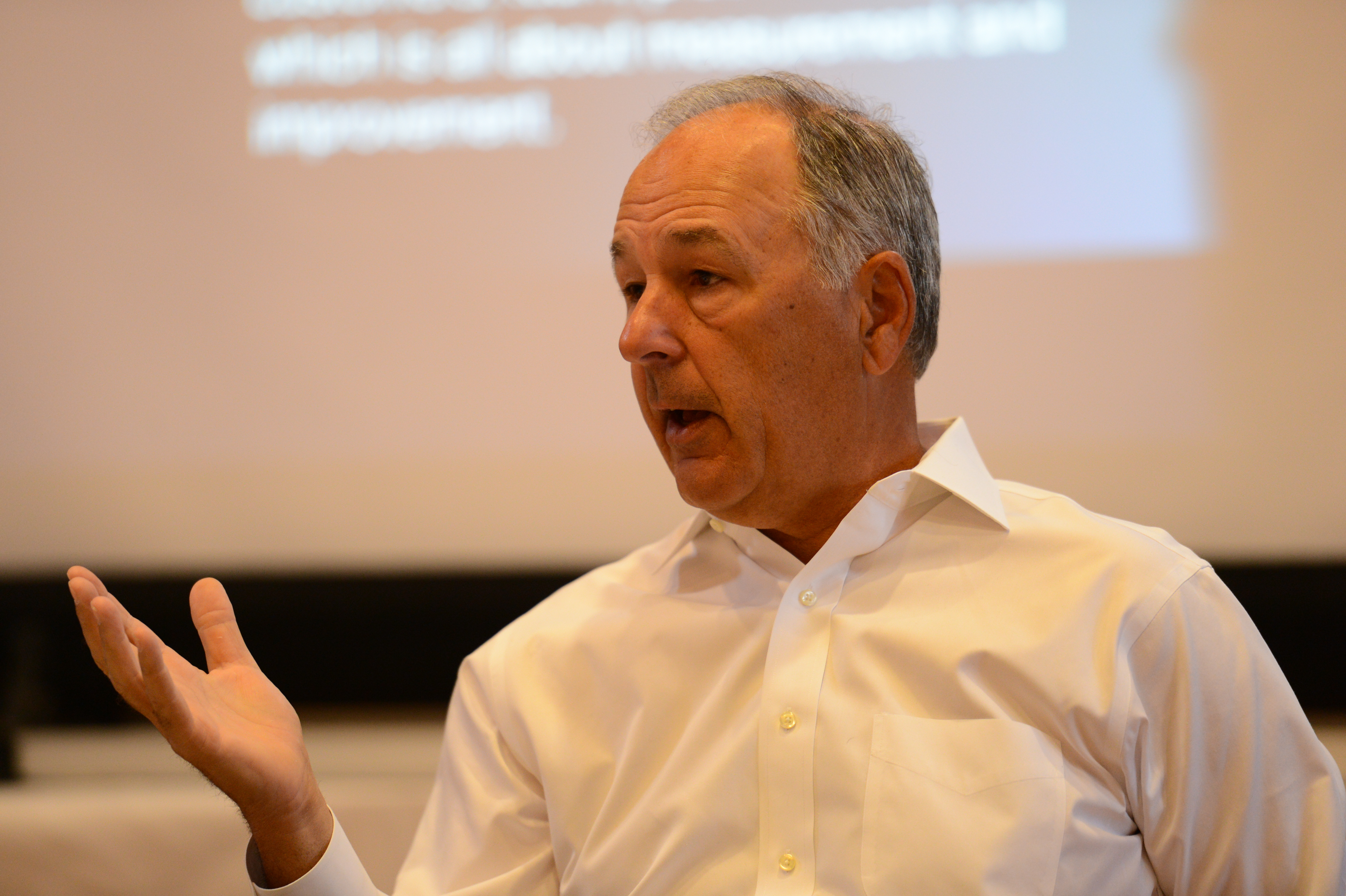A man in a white shirt speaks and gestures with his hand during a presentation.