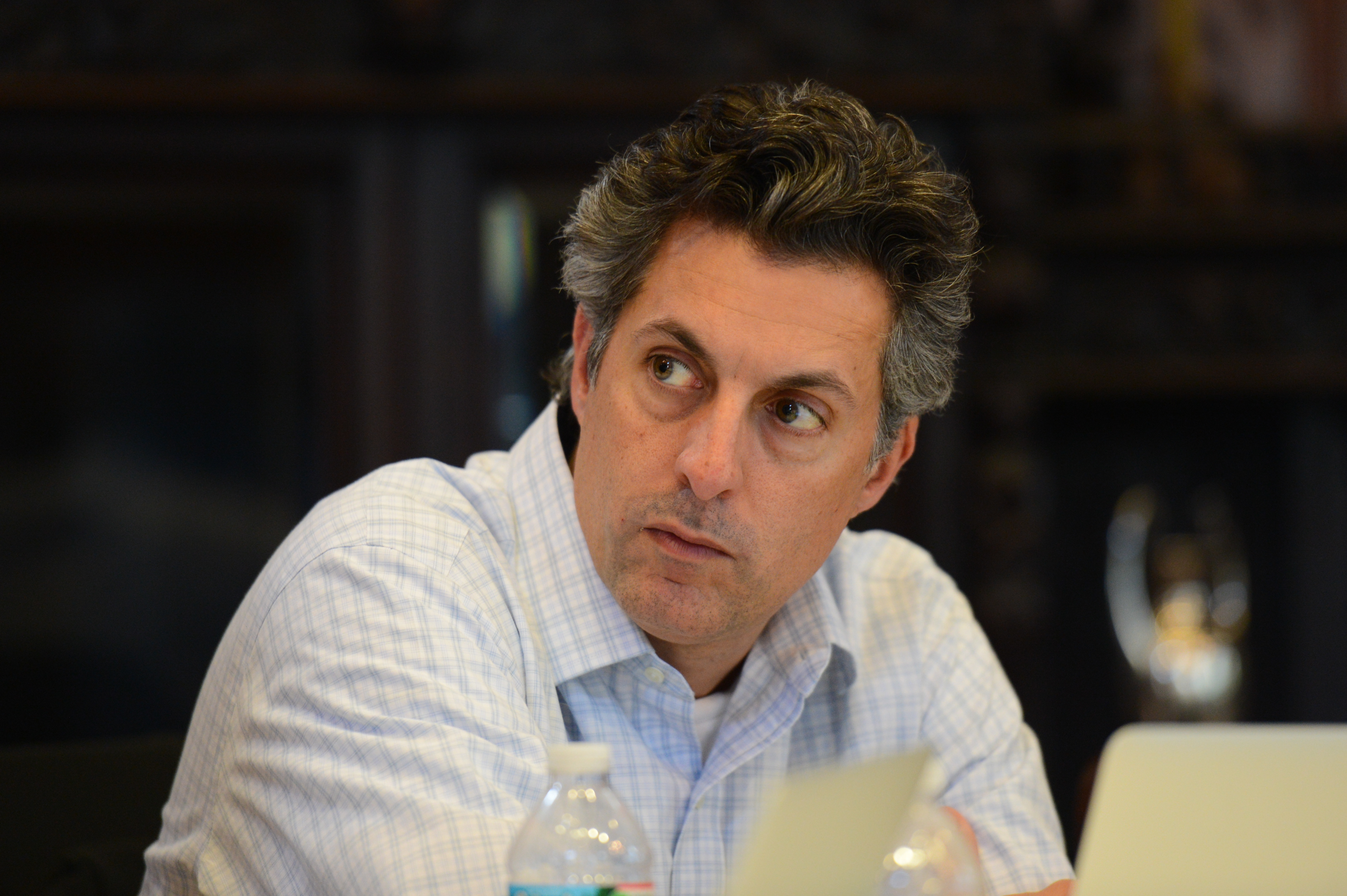 A man in a white shirt sits at a table, looking to the side with a focused expression.