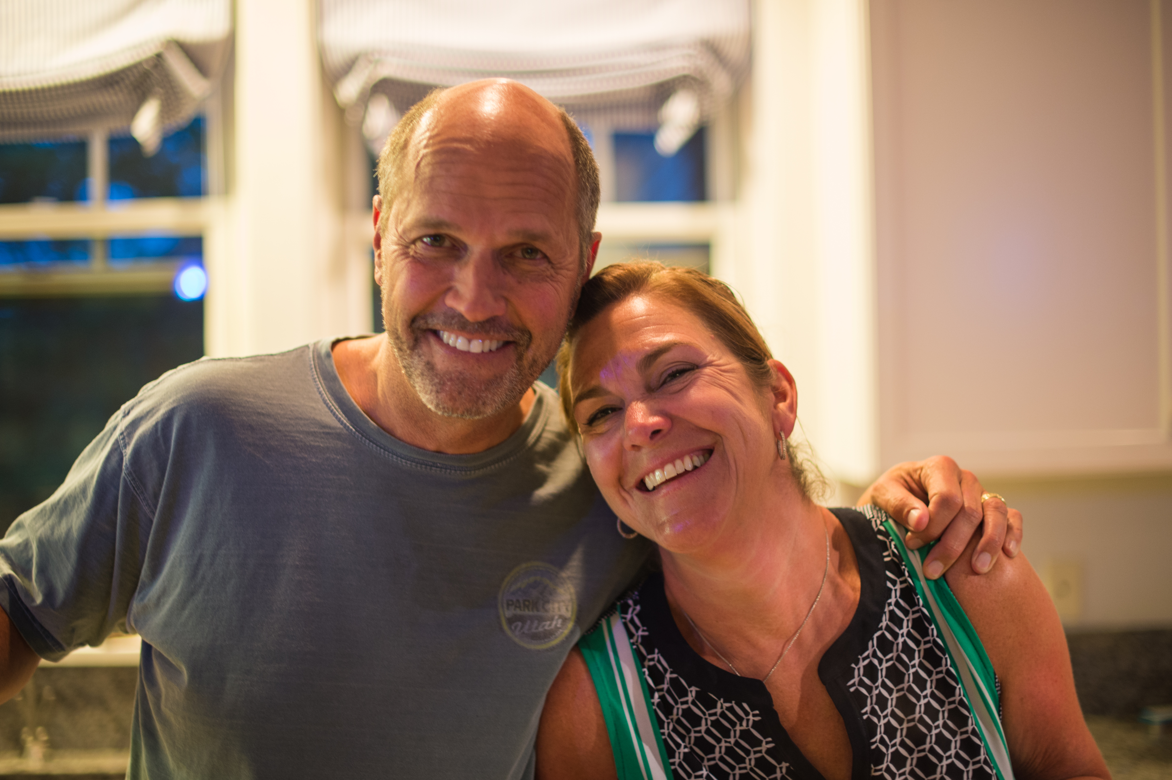A smiling man and woman pose together in a kitchen, with the man's arm around the woman's shoulder.