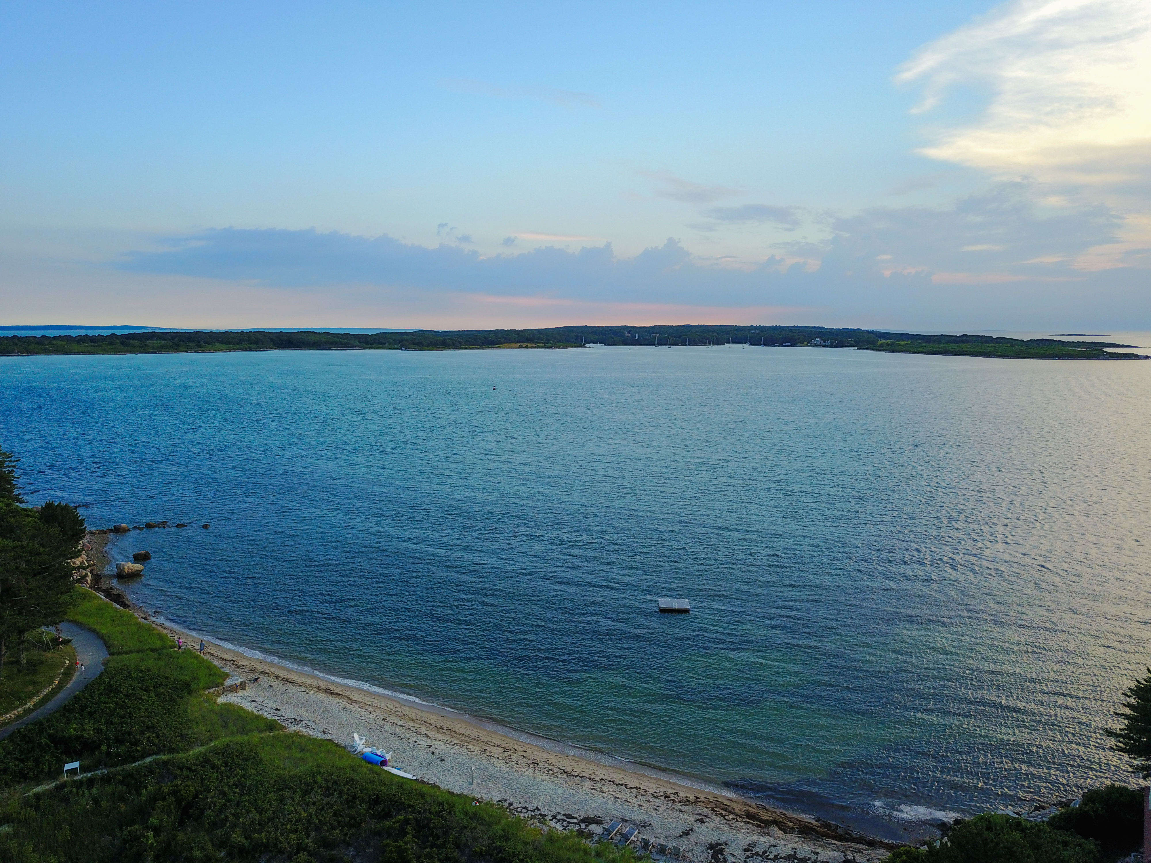 A calm coastal view with a sandy beach, blue water, a small floating dock, and distant land on the horizon.
