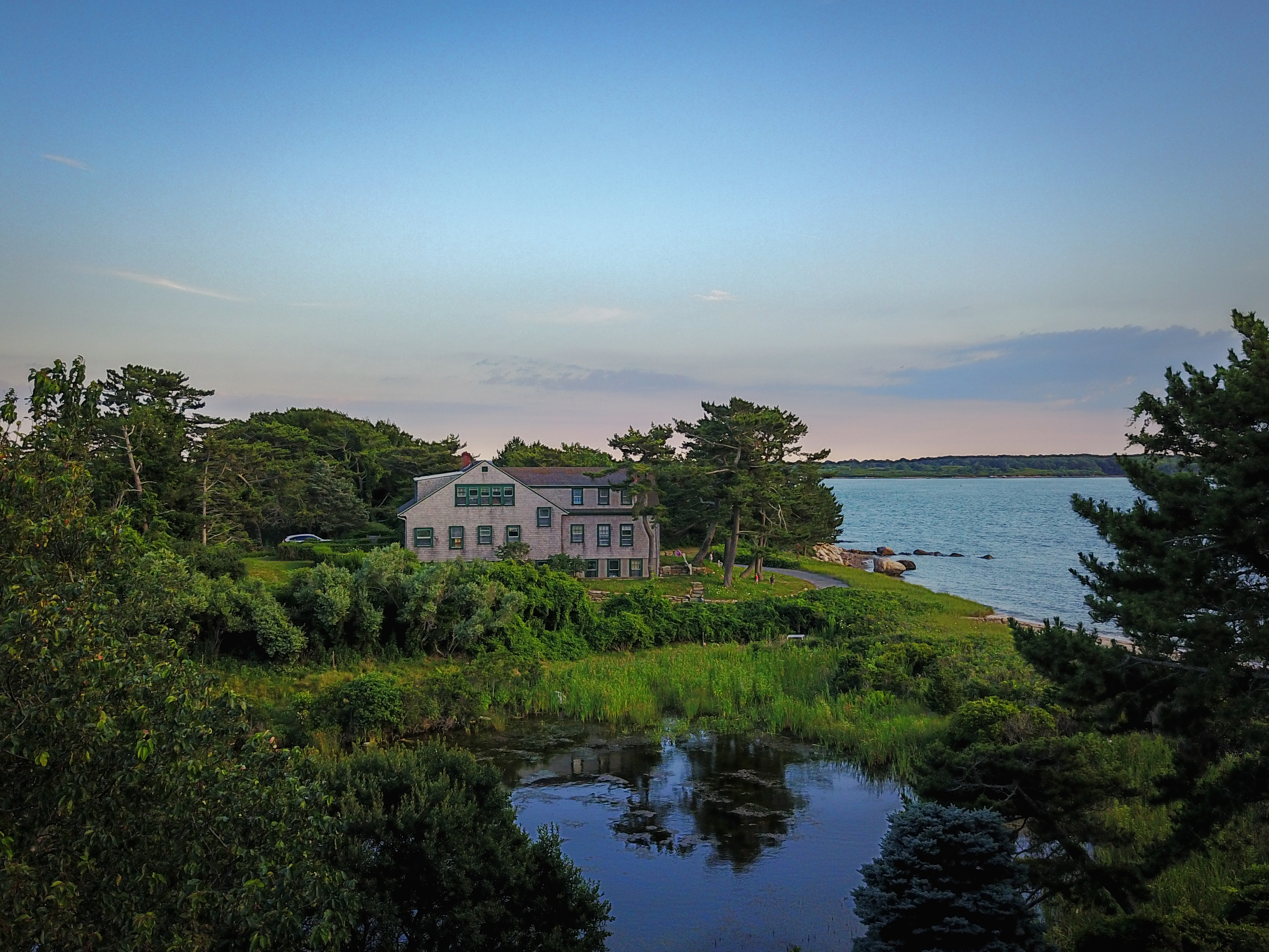 A large house surrounded by trees sits near the water, with a small pond in the foreground.
