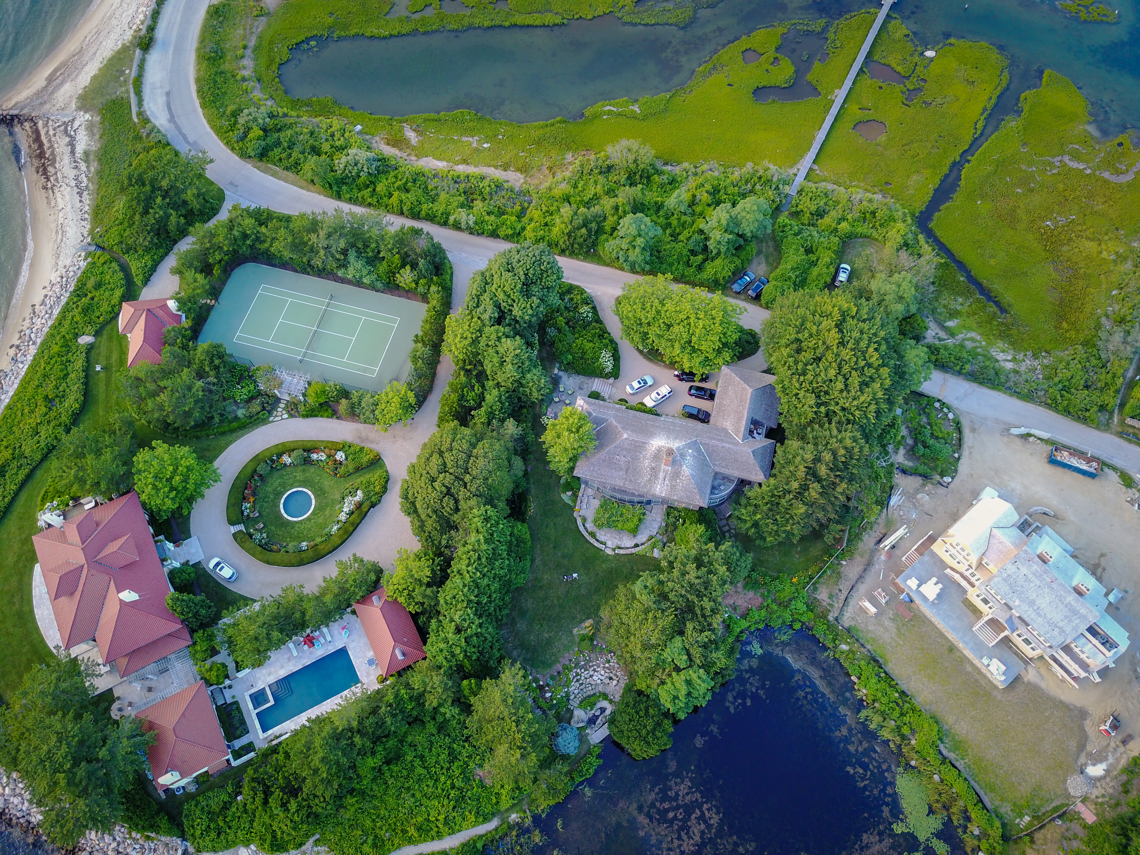 Aerial view of a coastal estate with houses, a tennis court, a swimming pool, and a pond surrounded by greenery.
