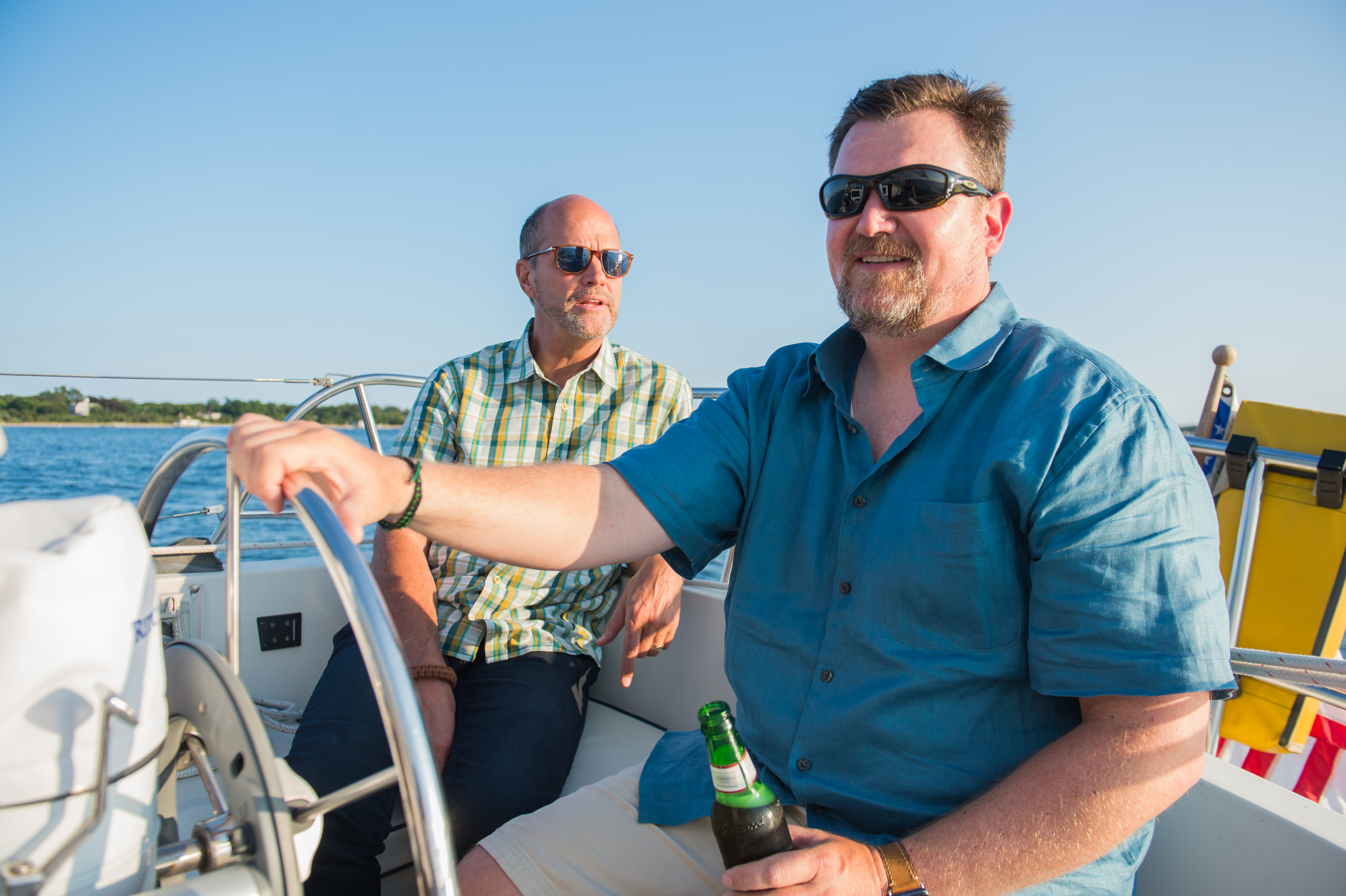 Two men on a sailboat, one steering and the other sitting, both wearing sunglasses and enjoying the ride.