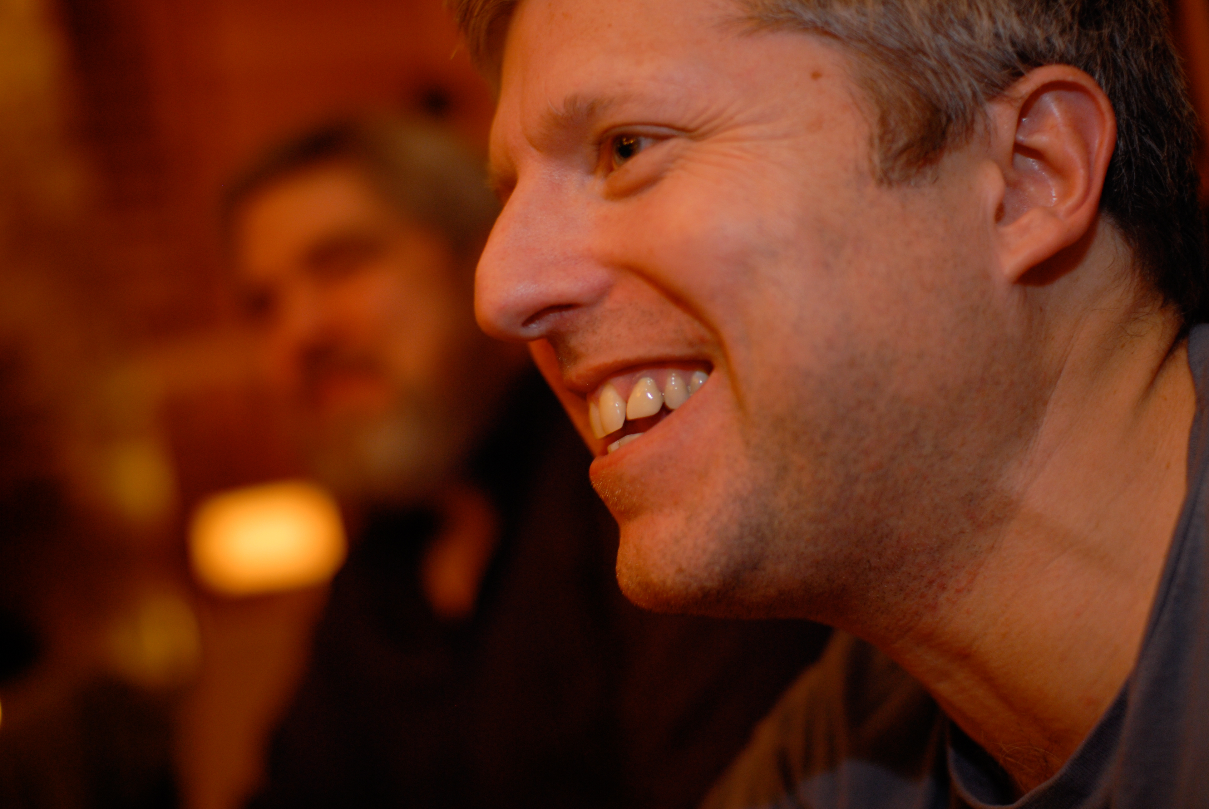 A man in the foreground is smiling, while another person is blurred in the background in a dimly lit setting.
