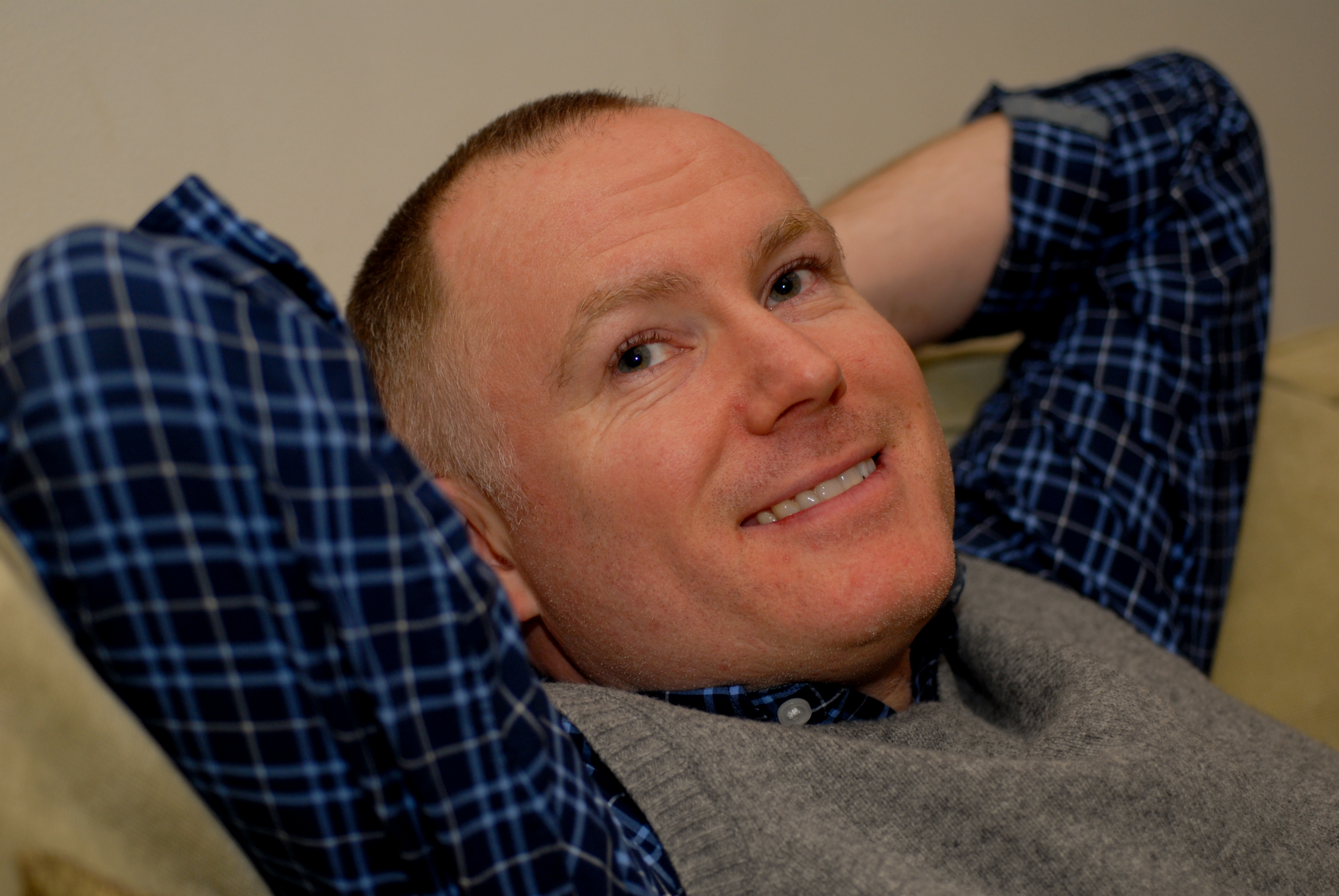 A man in a plaid shirt and vest leans back on a couch, smiling and relaxing.