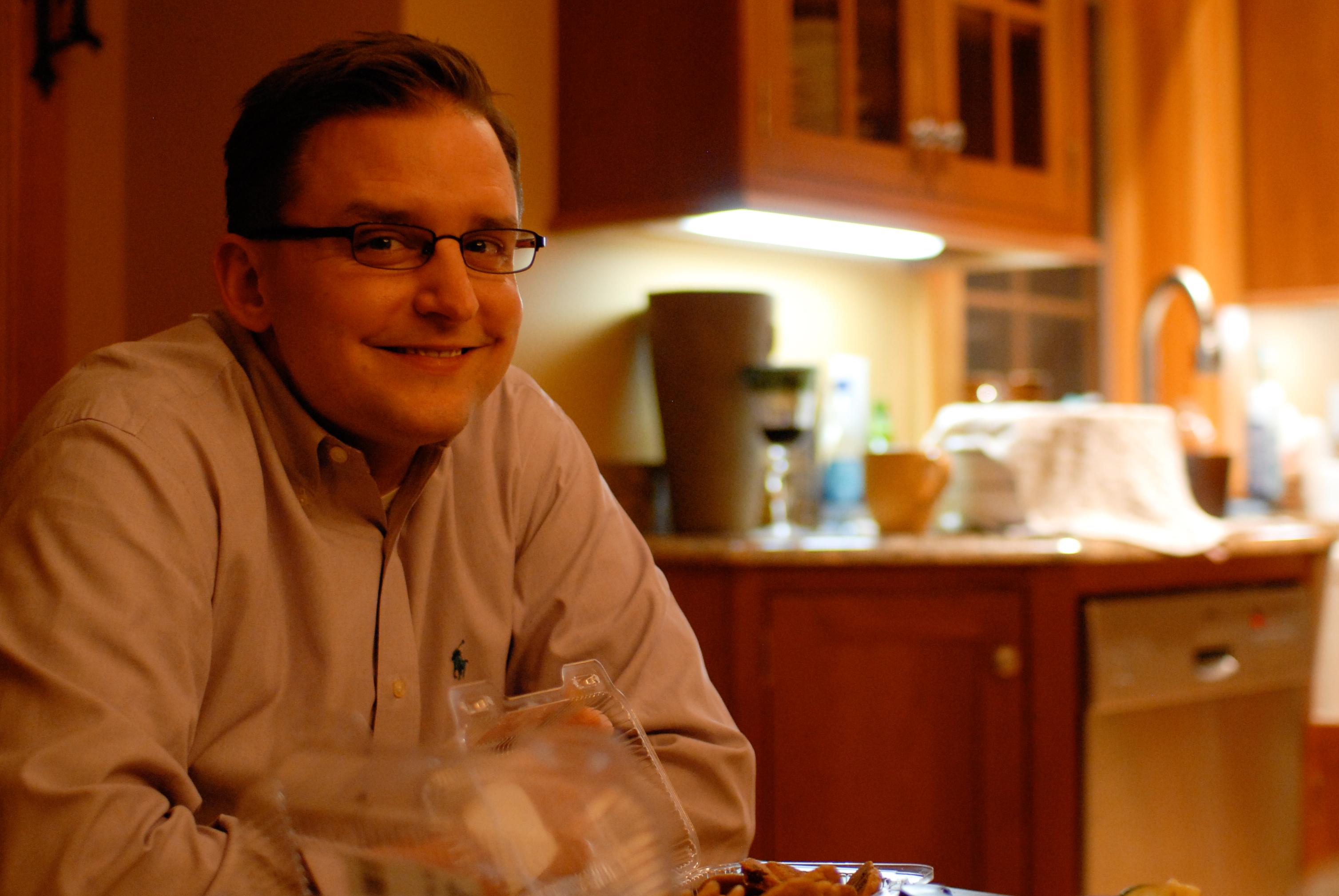 A man wearing glasses and a button-up shirt smiles while sitting at a table with food in a kitchen.