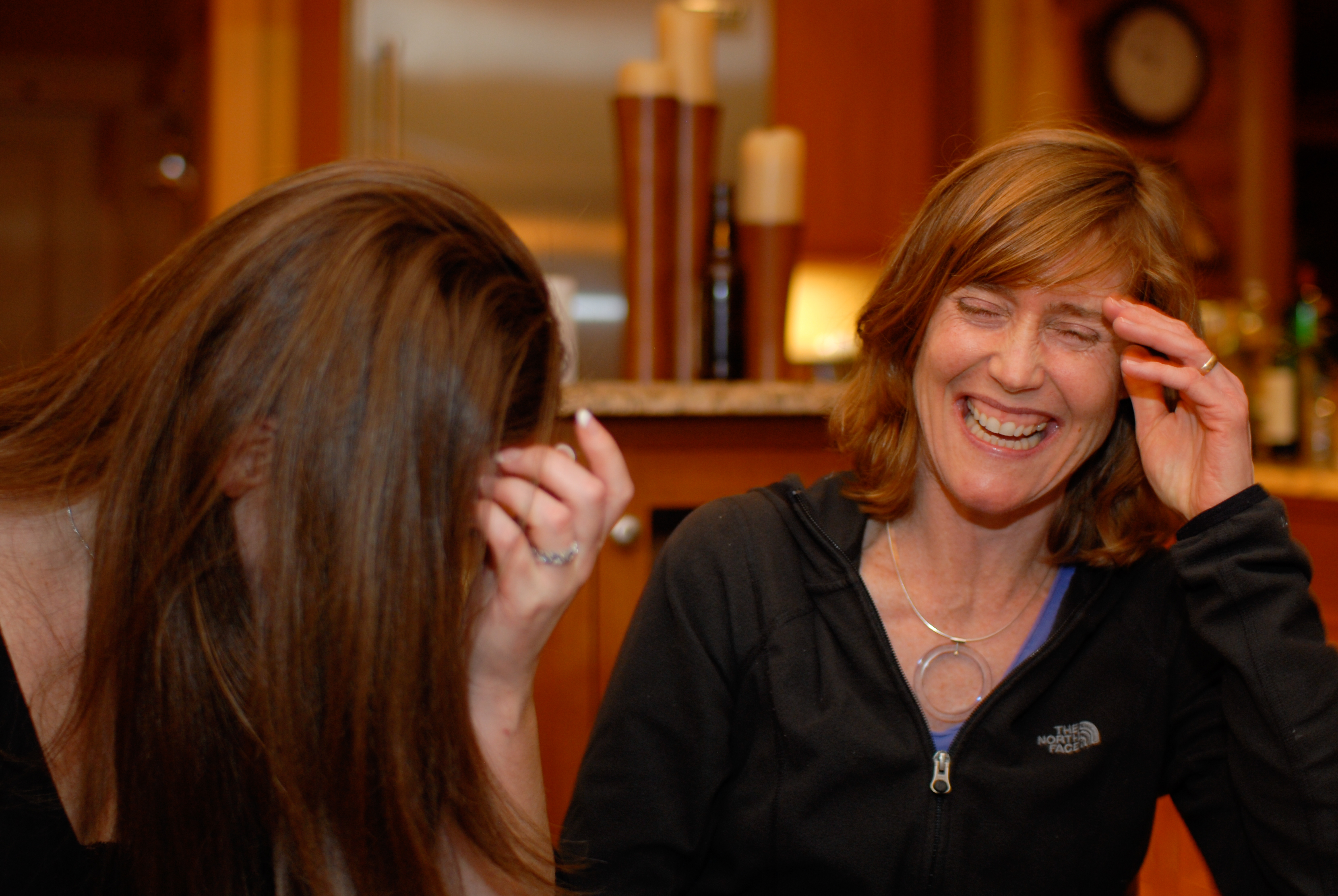 Two women laughing, one covering her face with her hand, in a warmly lit indoor setting.