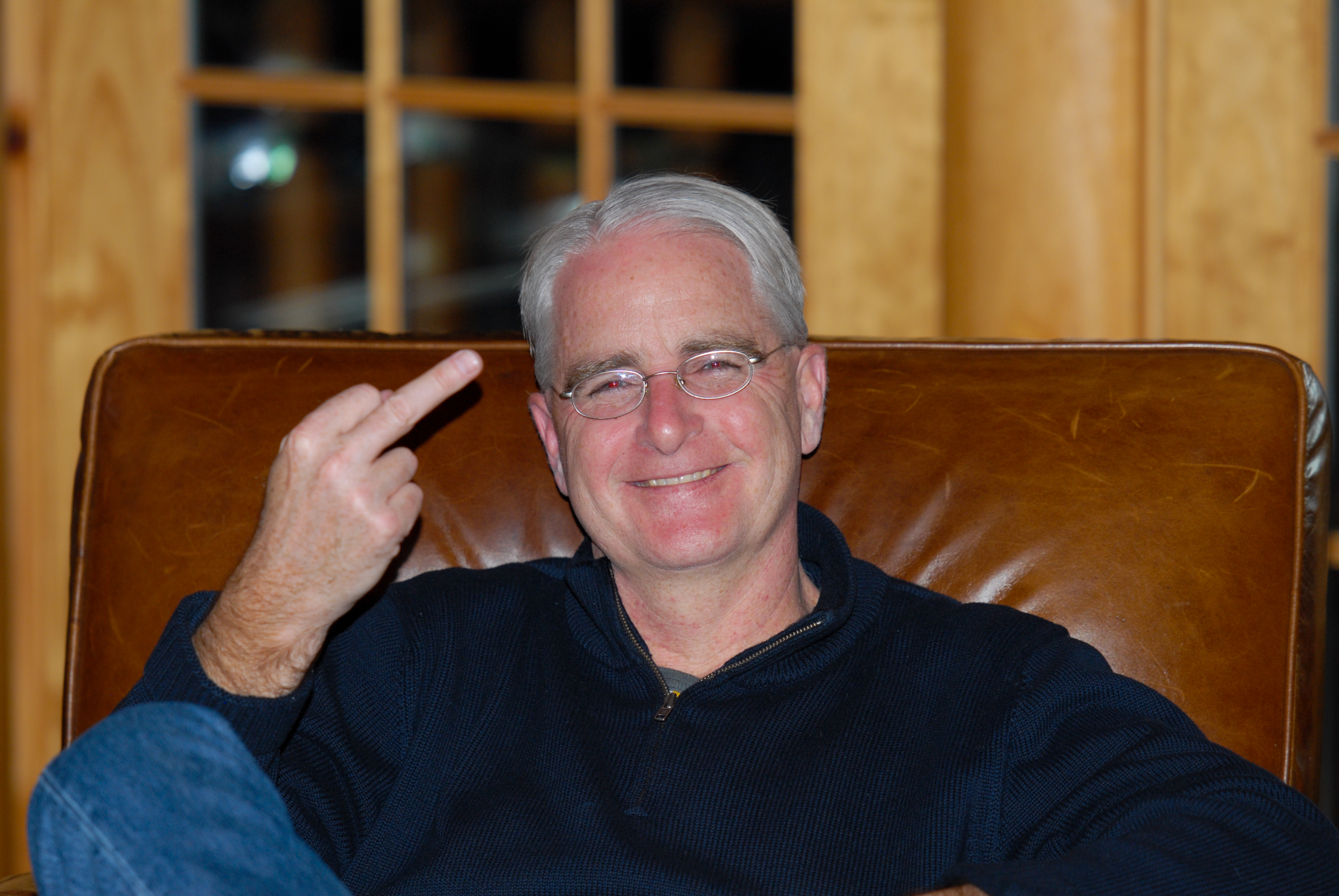A man wearing glasses and a dark sweater sits on a leather chair, smiling and raising his middle finger.